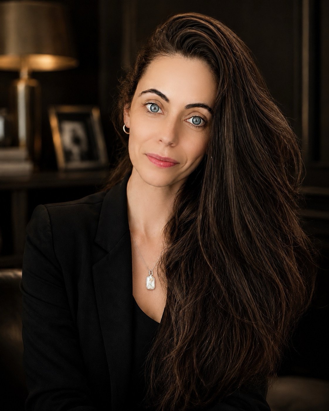 A woman with long dark hair, blue eyes, and fair skin, wearing a black blazer and a silver necklace with a rectangular pendant, posed indoors with a dark background and warm lighting.
