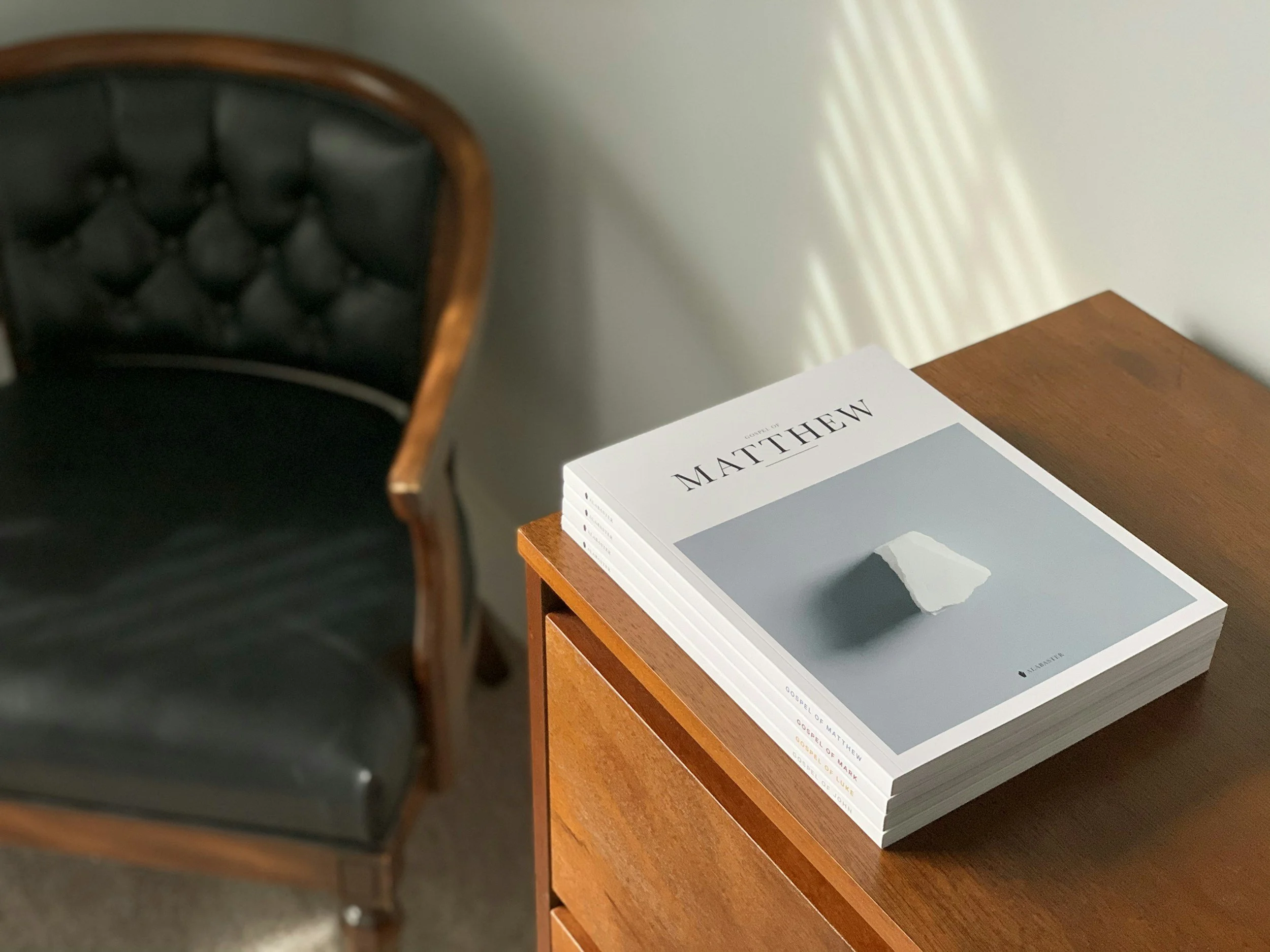 A black tufted leather chair with a wooden frame on the left side of the image. Next to it, on a wooden table, there is a stack of books with mostly white covers and black text, with some having gray and red accents. The top book has the title 'Gospel of Matthew'. The background features a plain light-colored wall with sunlight creating diagonal lines of light and shadow.