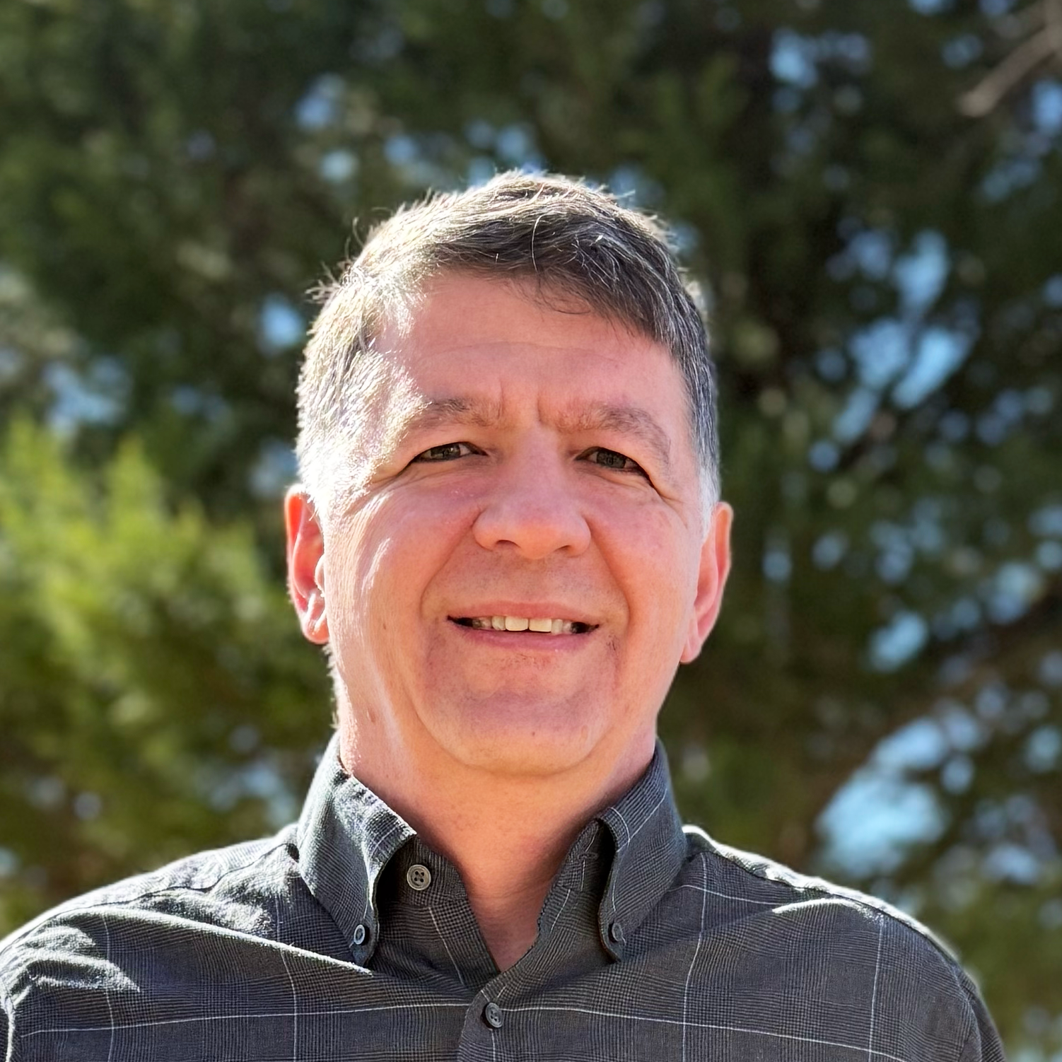 A smiling middle-aged man with short hair wearing a gray plaid shirt outdoors with trees in the background.