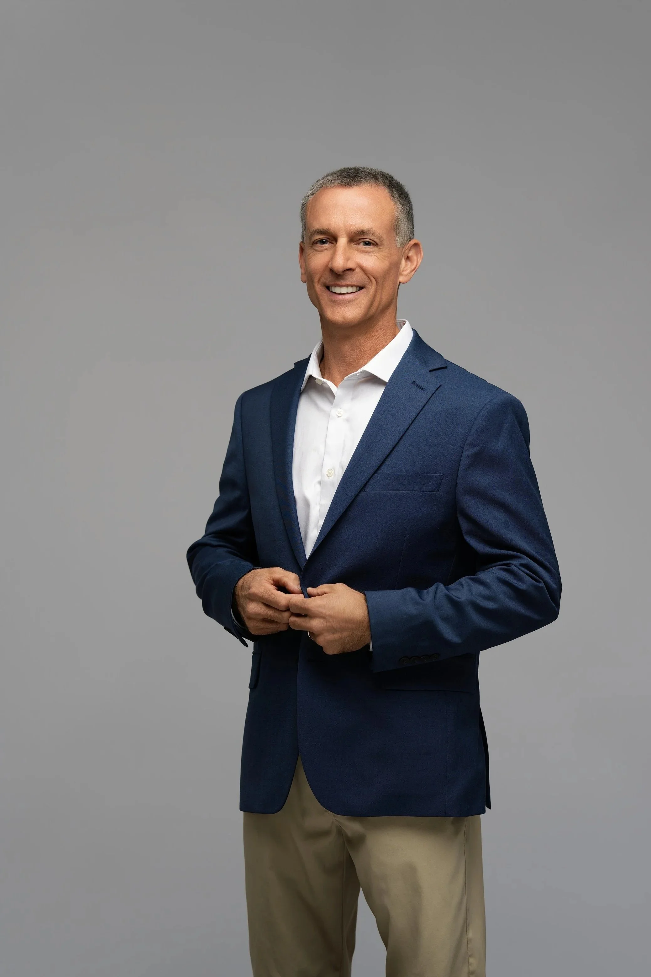 Smiling middle-aged man in a navy blazer and beige pants standing against a plain gray background.