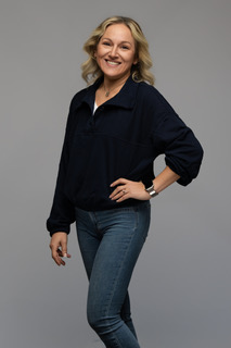 Smiling woman with blonde hair and a denim shirt standing in front of a plain gray background.