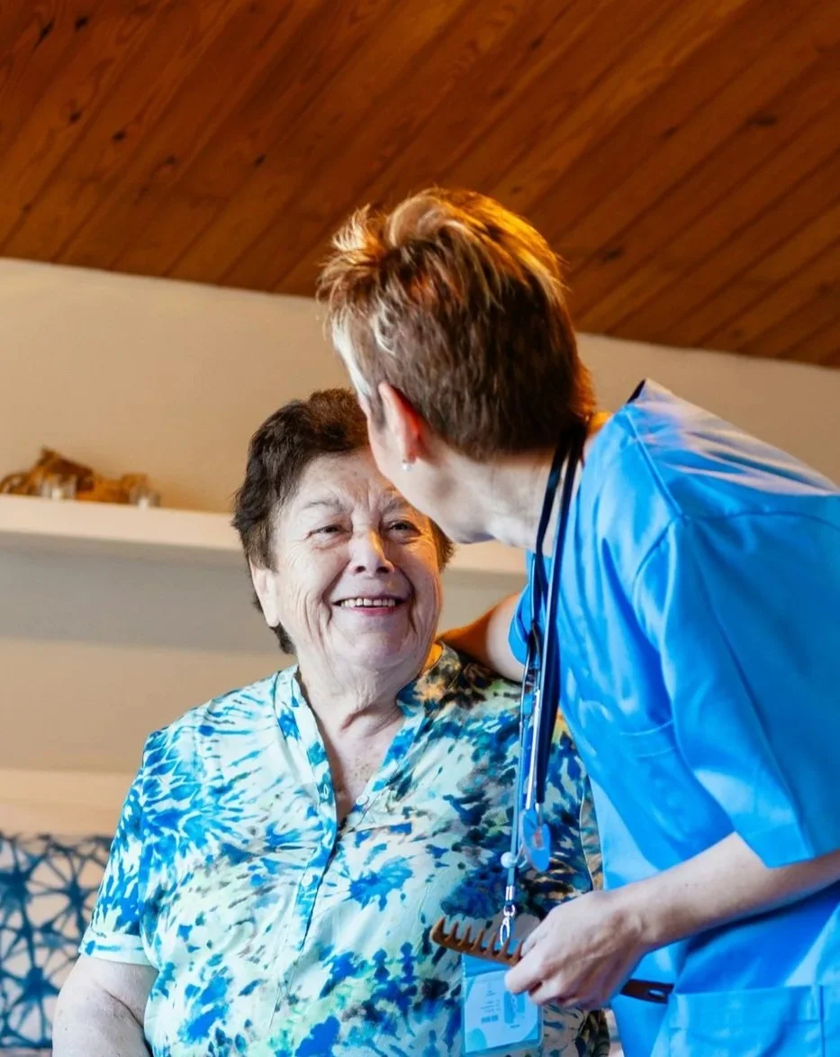 A healthcare worker wearing blue scrubs is smiling and interacting with an elderly woman. The woman has short, dark hair and is wearing a patterned blue and white blouse. They are indoors, in a room with wooden ceiling and shelves.