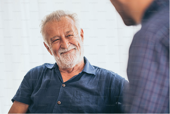 An elderly man with white hair and a beard smiling while talking to a younger man in a checkered shirt.