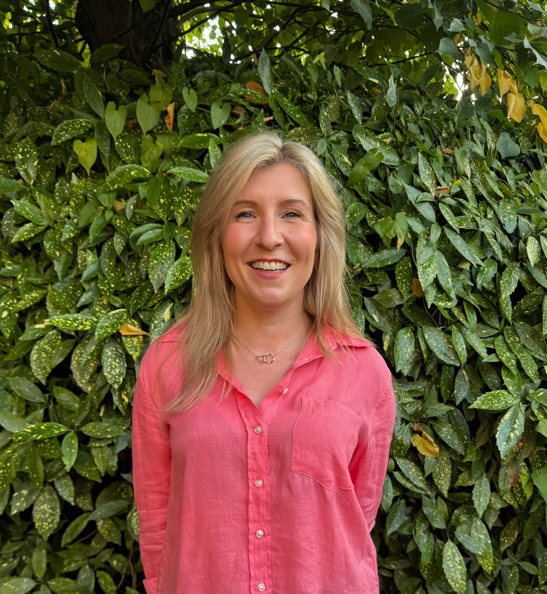 A smiling woman with blonde hair wearing a pink shirt standing in front of green leafy shrubbery.