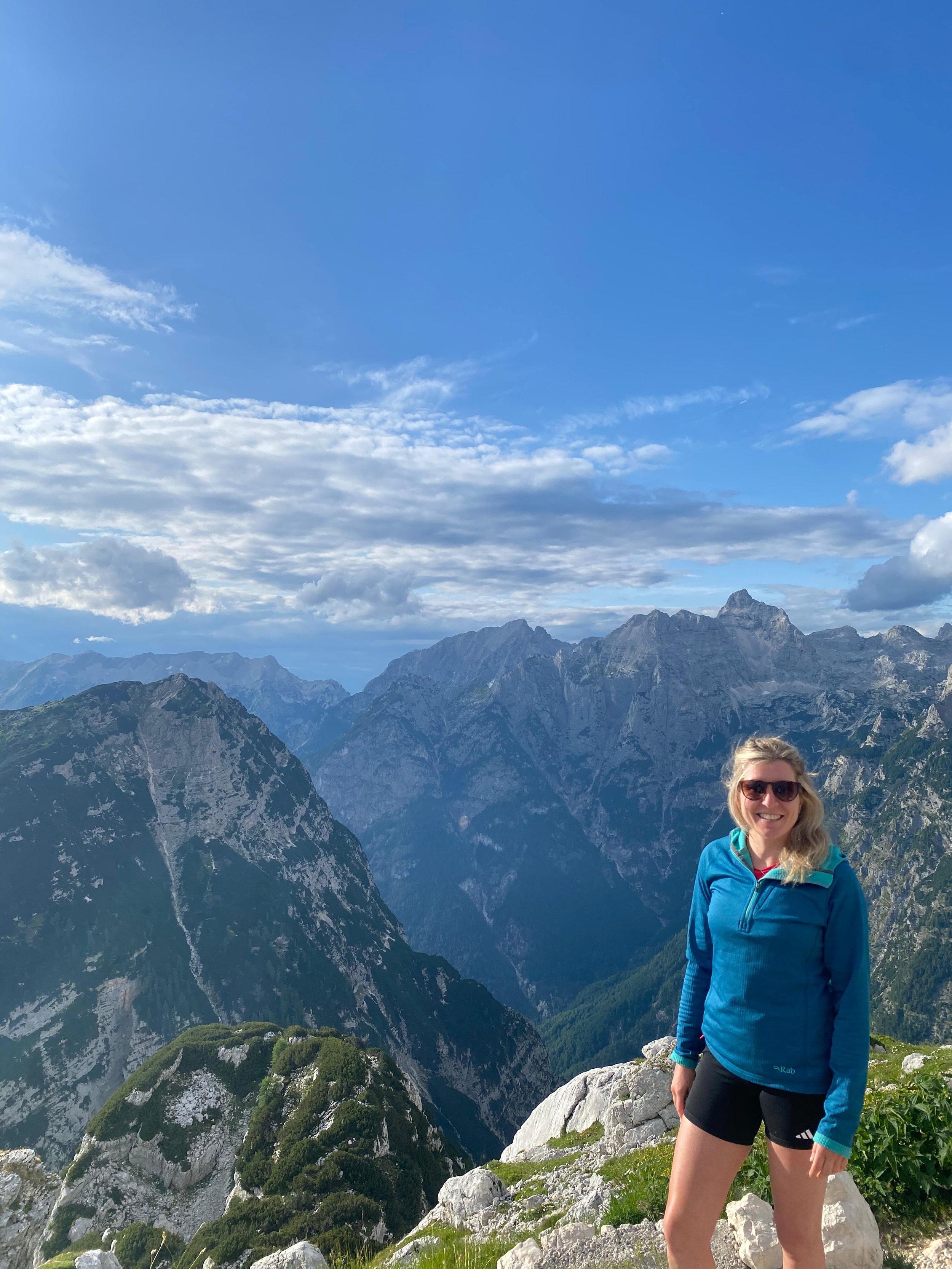 A woman smiling in a blue jacket and black shorts, standing on a rocky ledge in a mountainous area with rugged peaks, green slopes, and a blue sky with scattered clouds in the background.
