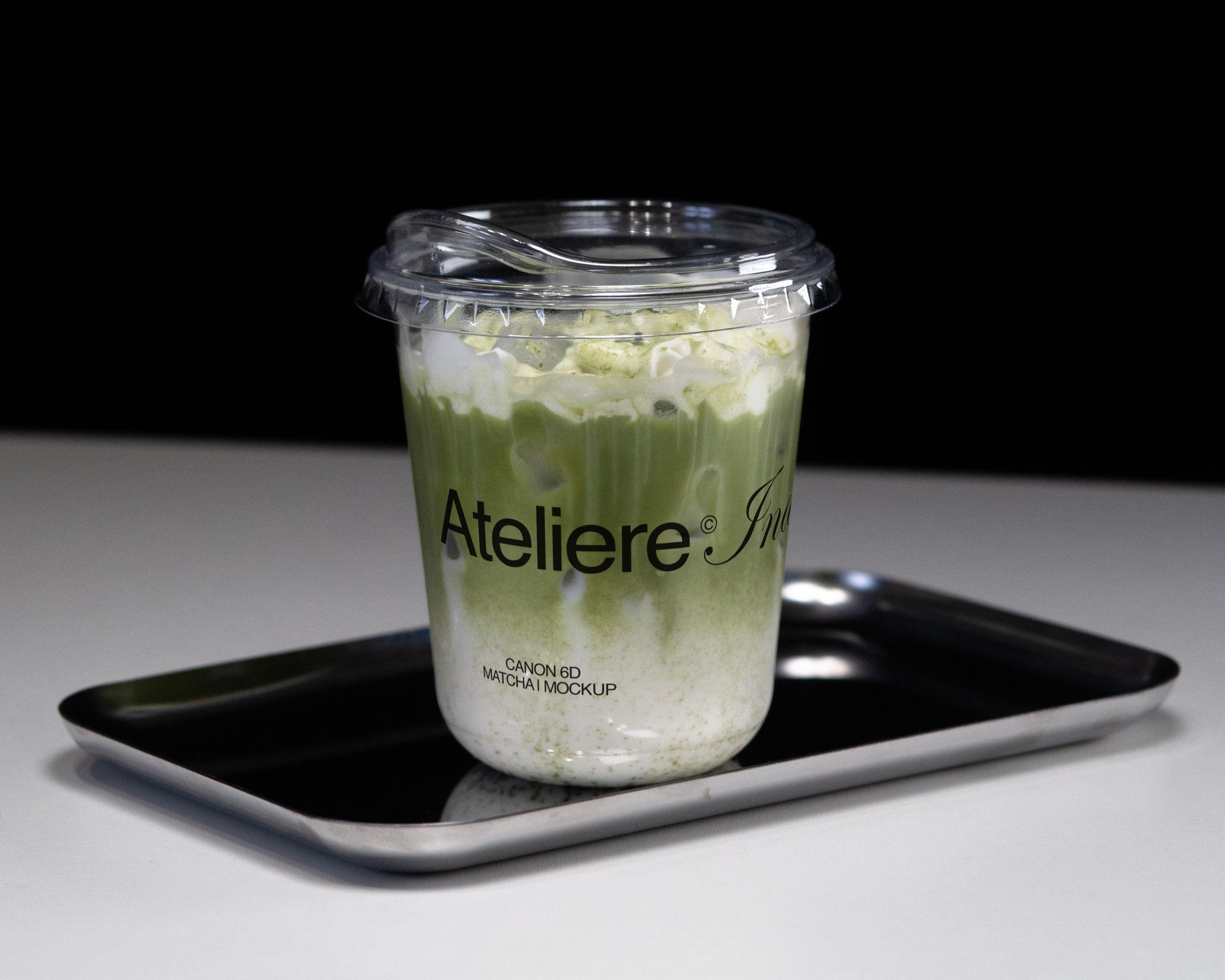 A plastic cup filled with a green drink topped with whipped cream and matcha powder, placed on a black tray on a white surface against a black background.