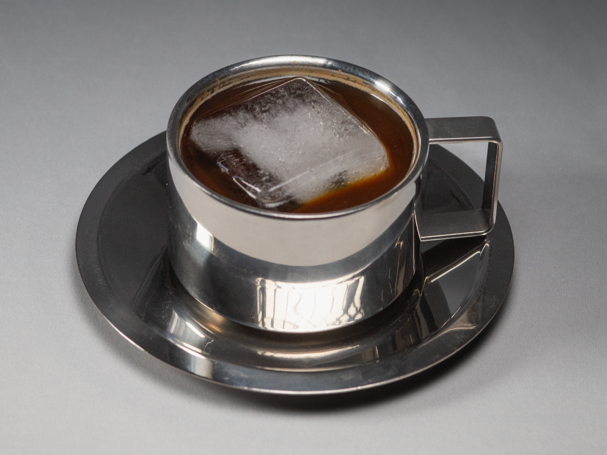 A metal cup filled with iced coffee and a large ice cube, placed on a metal saucer on a gray surface.