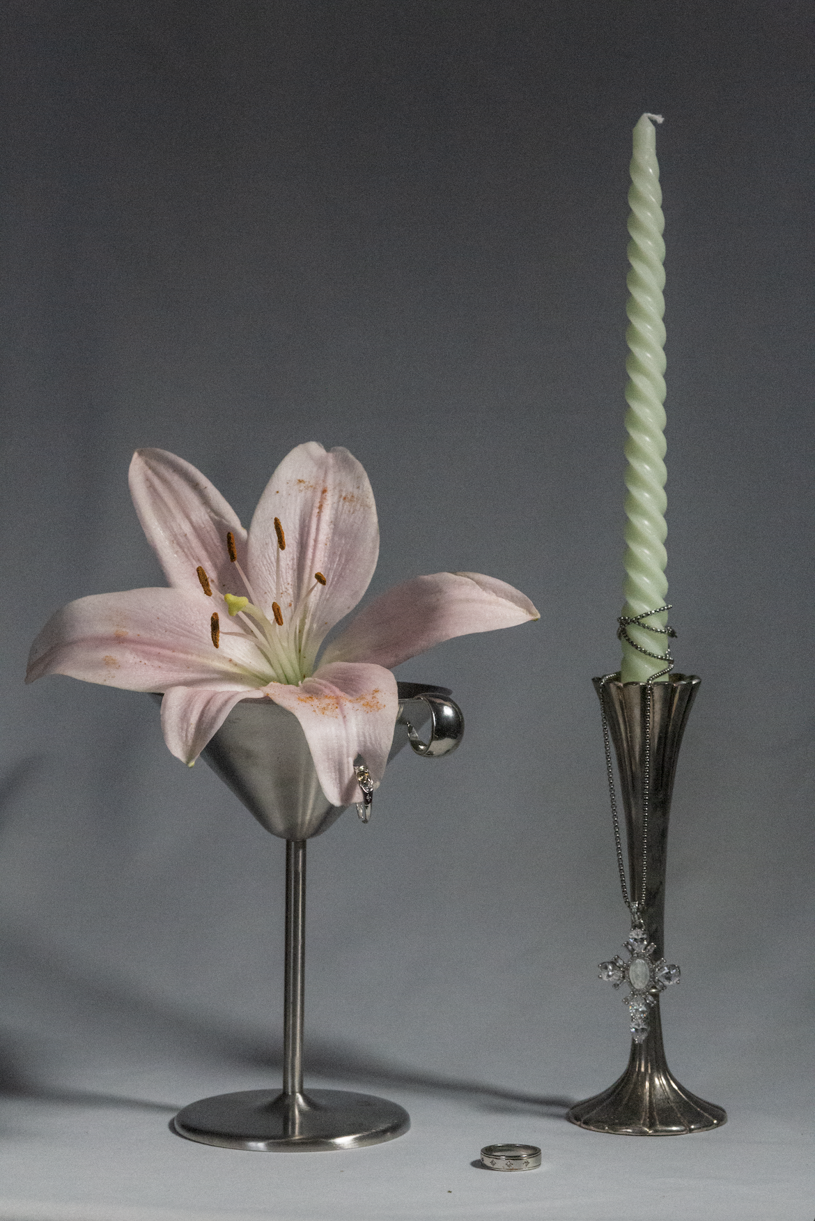 A pink lily flower and a twisted white candle in a silver holder with jewelry, including rings and a cross necklace, on a gray background.