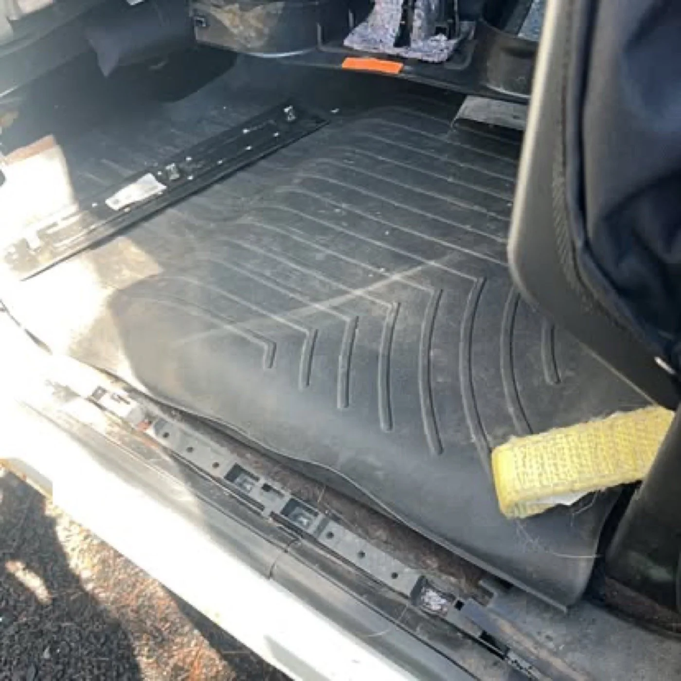 Inside a vehicle, showing a black rubber floor mat with grooves, a yellow cloth on the right side, and part of a black seat on the right. The vehicle appears to be in the process of being cleaned or repaired.