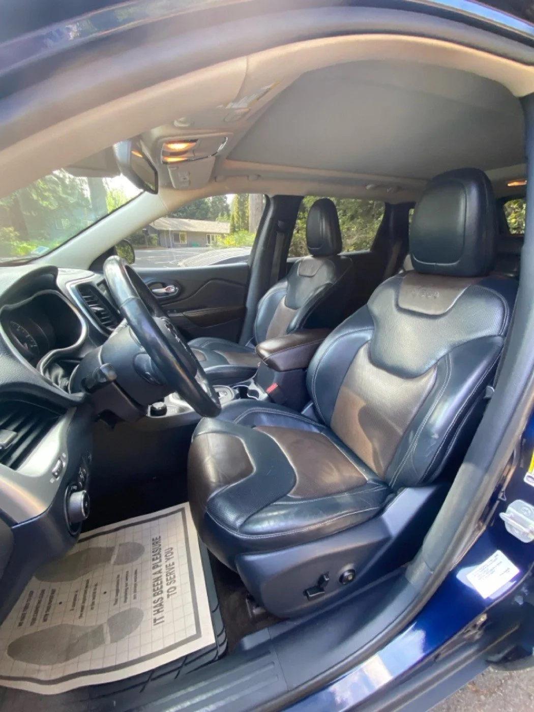 The interior of a vehicle, showing the front passenger and driver seats with leather upholstery, a steering wheel, dashboard, and overhead console with a light, taken on a sunny day with trees visible outside.