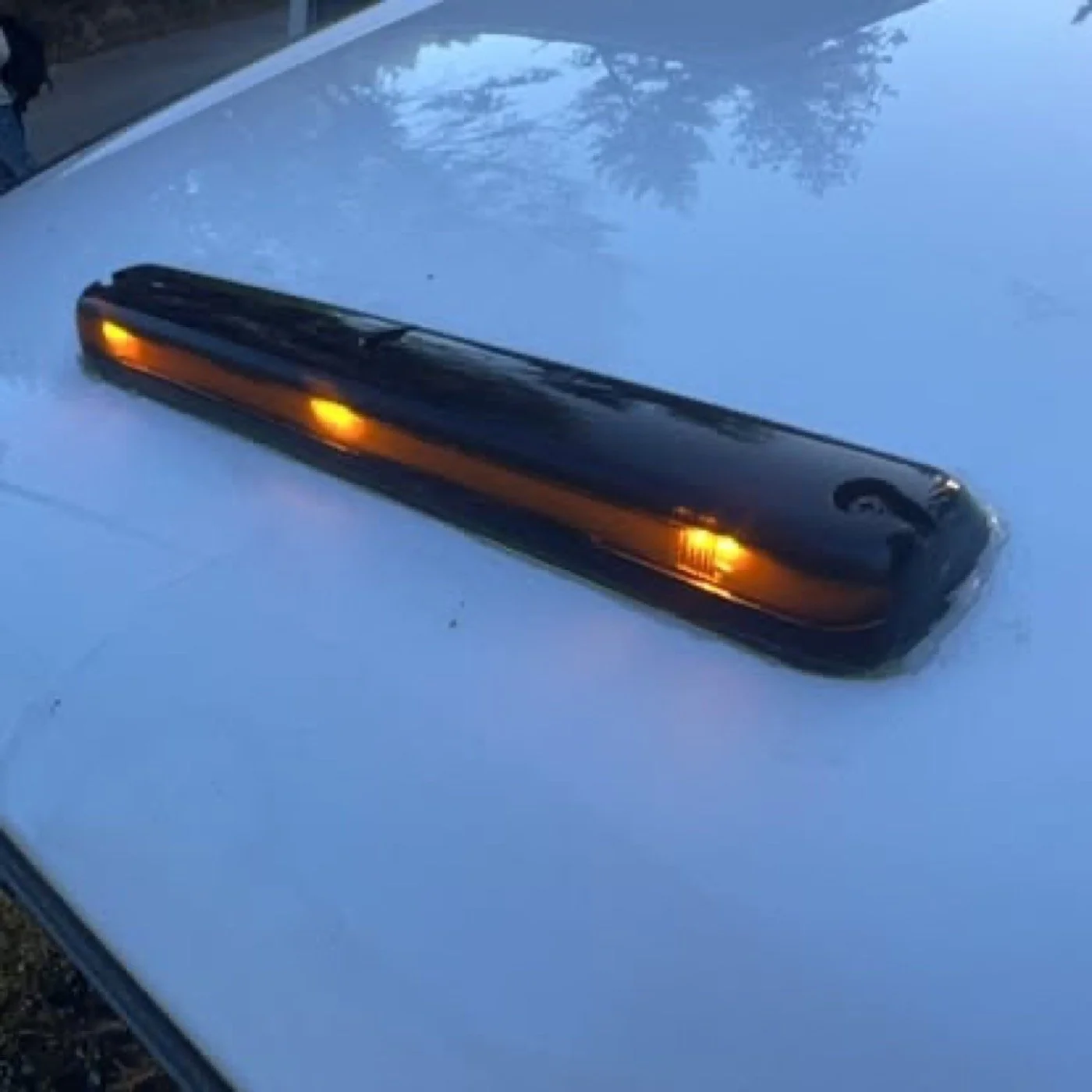 Close-up of a car's side mirror with the turn signal lights illuminated, reflecting trees and sky in the mirror, with a blue car roof in the background.