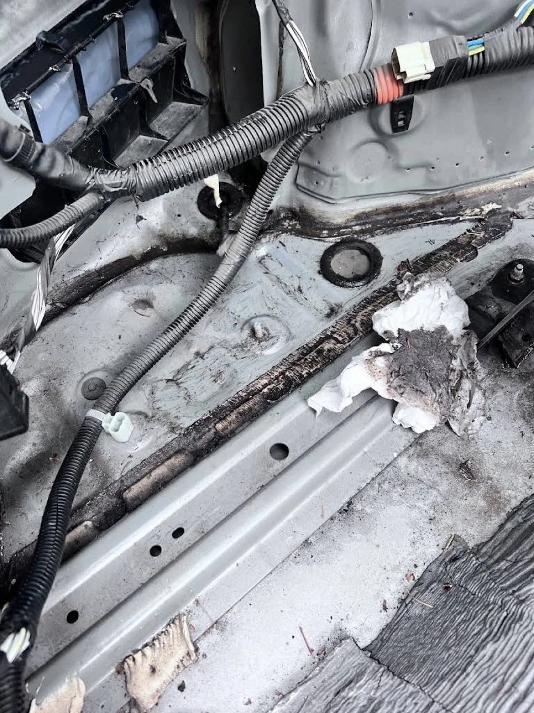Close-up of a vehicle's interior showing exposed wires, a rusty metal surface, and dirty paper towels on a floor with worn wood.
