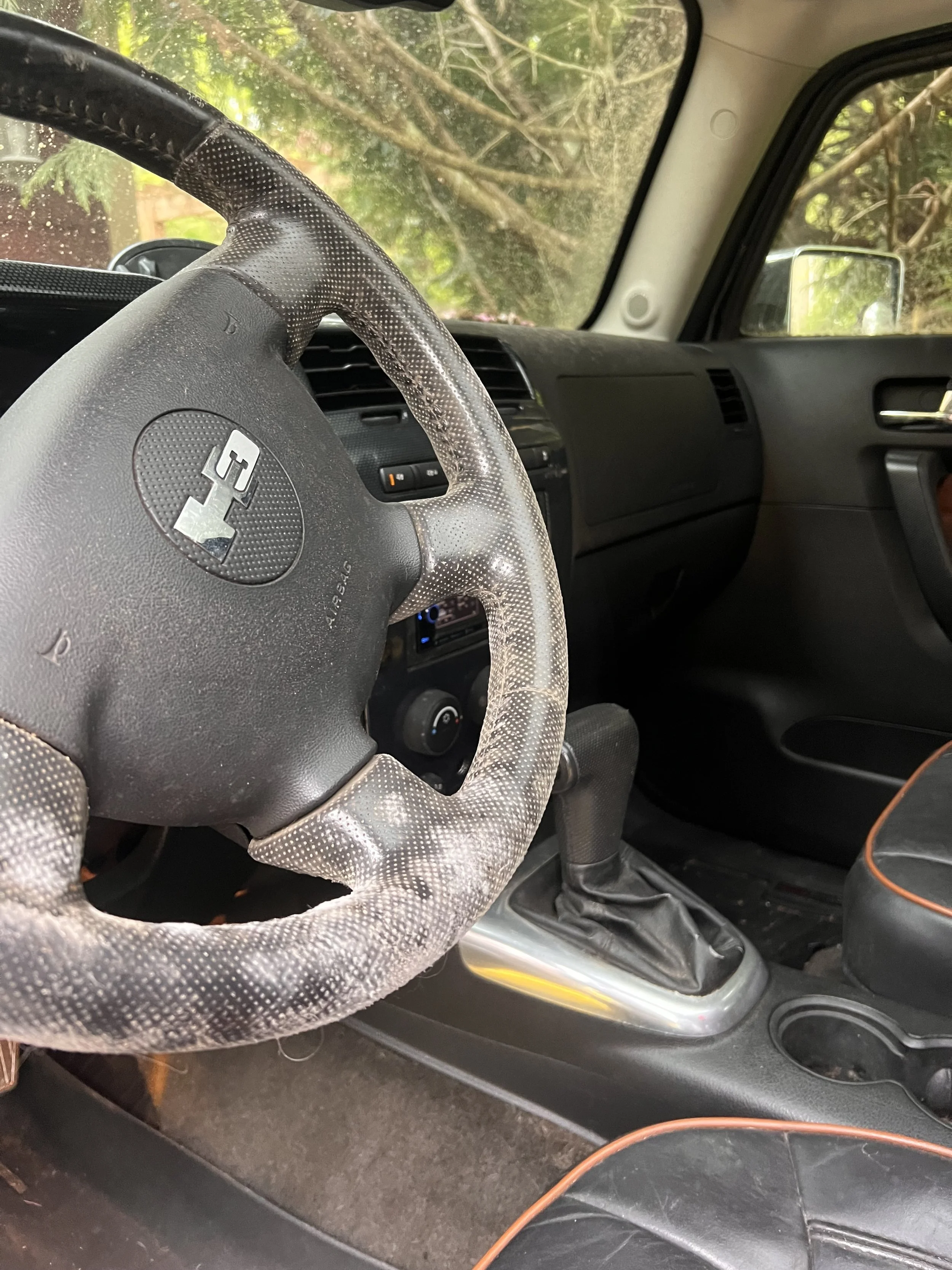 The interior of a vehicle showing a steering wheel with a faded textured cover, a dashboard with controls, and a gear shift. The window reveals foliage outside.