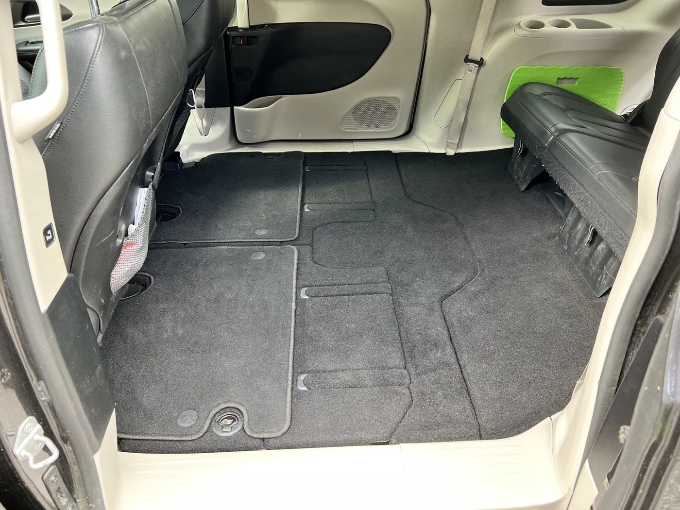 The interior of a vehicle showing the back seat area with black carpeted floors, a bench seat on the right, and the back of the front seats with storage pockets. The door panel includes a small speaker and storage compartment.