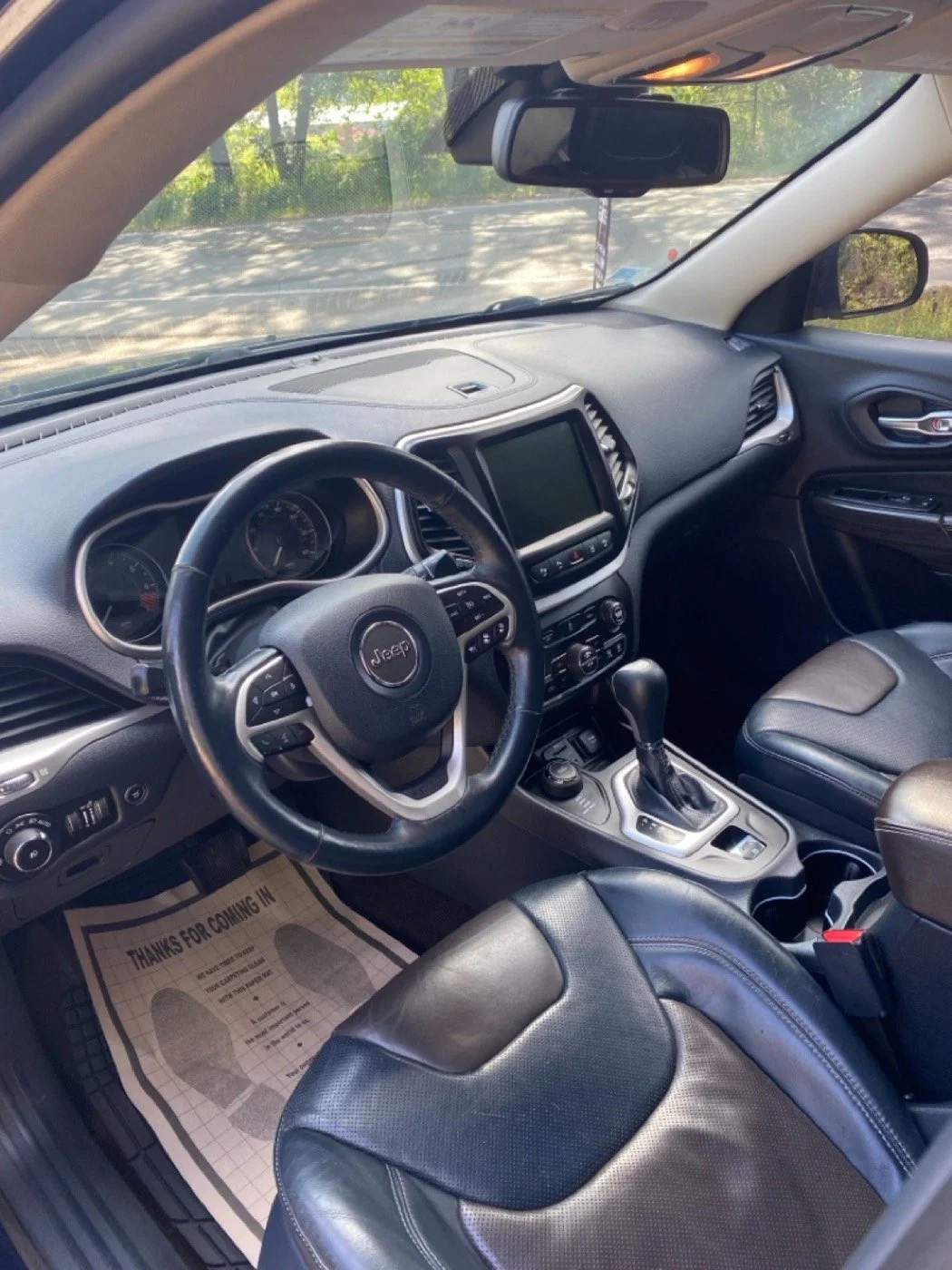 Interior view of a Jeep vehicle, showing the dashboard, steering wheel, center console with gear shift, and front passenger seat, with trees visible through the windshield.