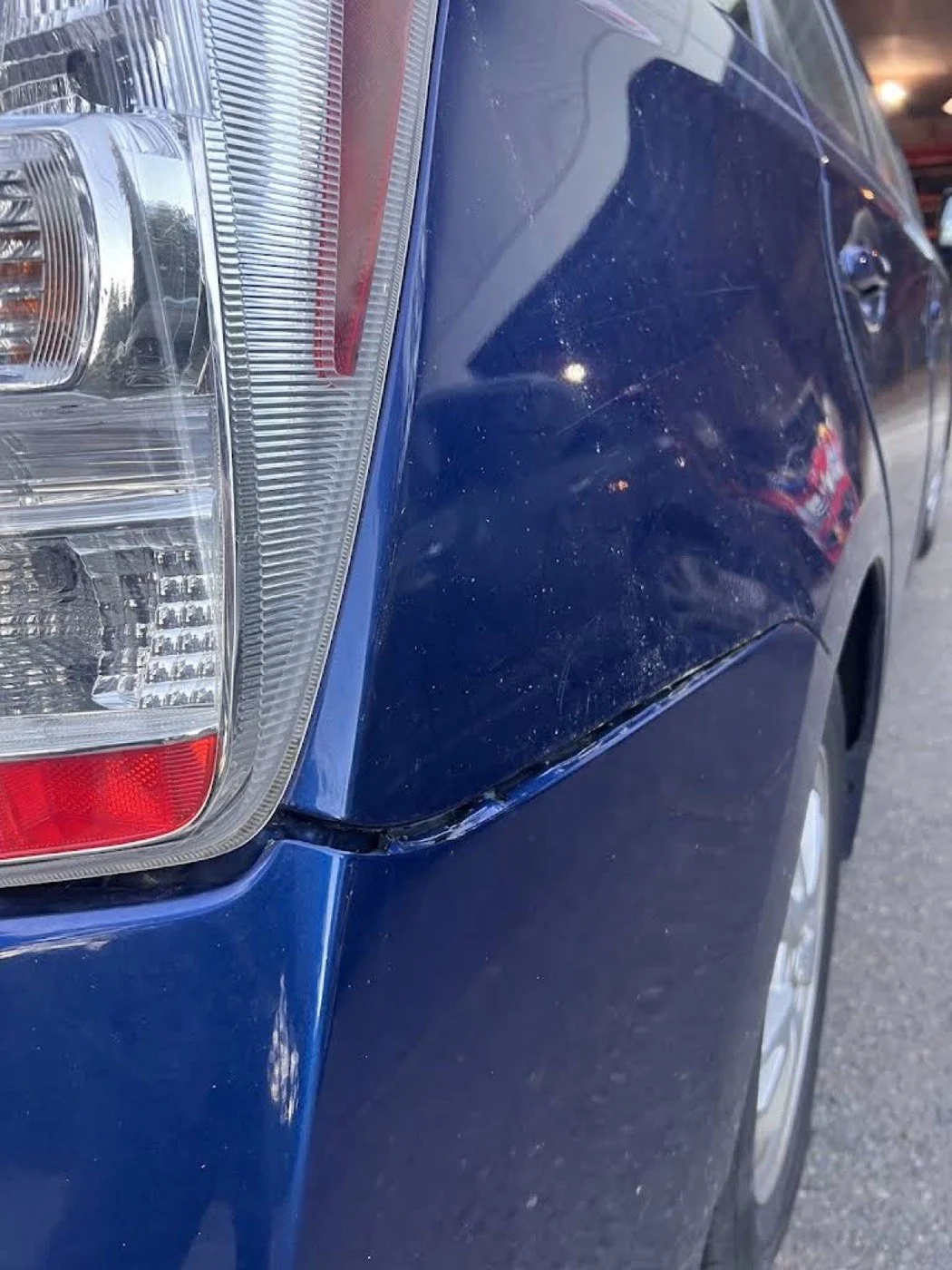 Close-up of the rear left side of a blue car with visible dirt and scratches, focusing on the taillight and fender.