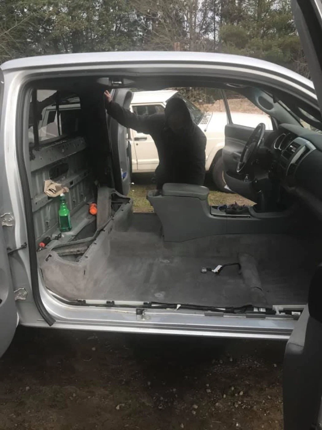 Interior of a vehicle with the driver side door open, showing a person dressed in black climbing into the vehicle, with its interior mostly empty and some objects on the floor and dashboard.