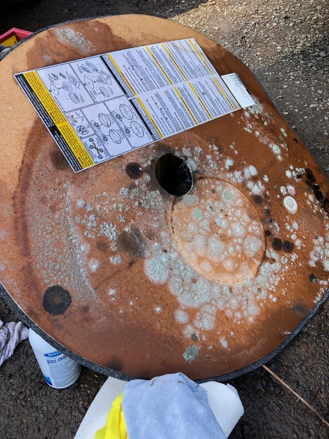 A rusty metal circular object, possibly a large disc or part of machinery, with white and greenish mold or lichen growth. On top of the object is a sheet of paper with instructions and diagrams, and a nearby spray bottle of glass cleaner on the groun