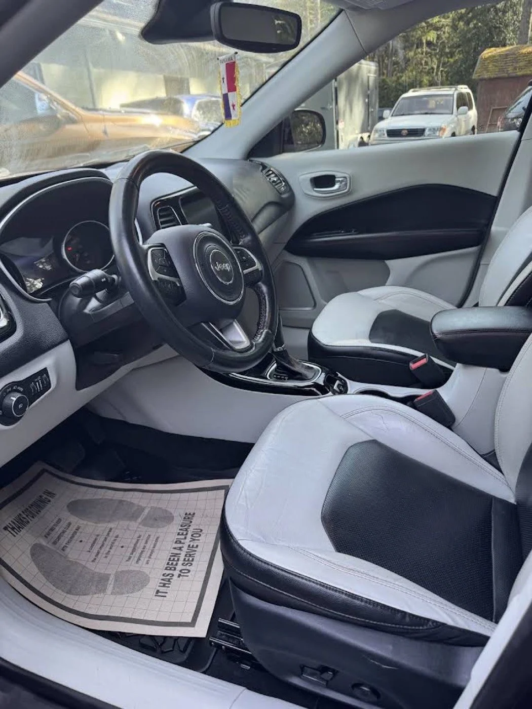 Interior of a Jeep vehicle showing the driver's seat, steering wheel, center console, and front passenger seat. The seats are white with black accents. There is a paper floor mat that reads, "It has been a pleasure to serve you." outside the vehicle,