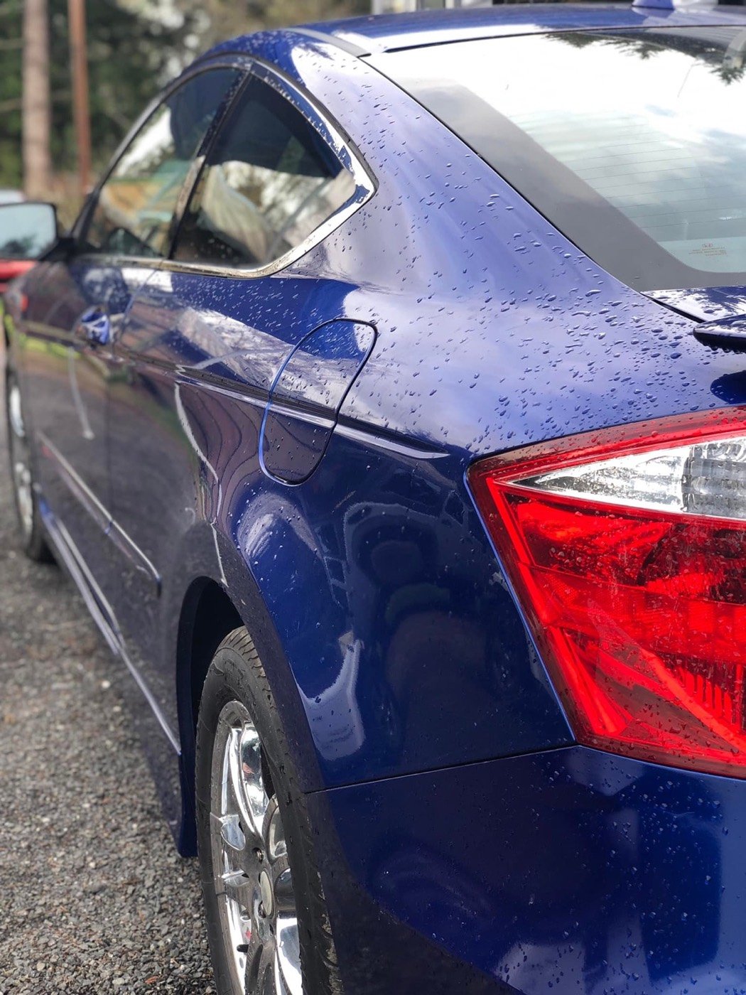 Close-up of a blue car with water droplets on the surface parked outdoors, showing the rear quarter panel, rear windshield, and part of the taillight.