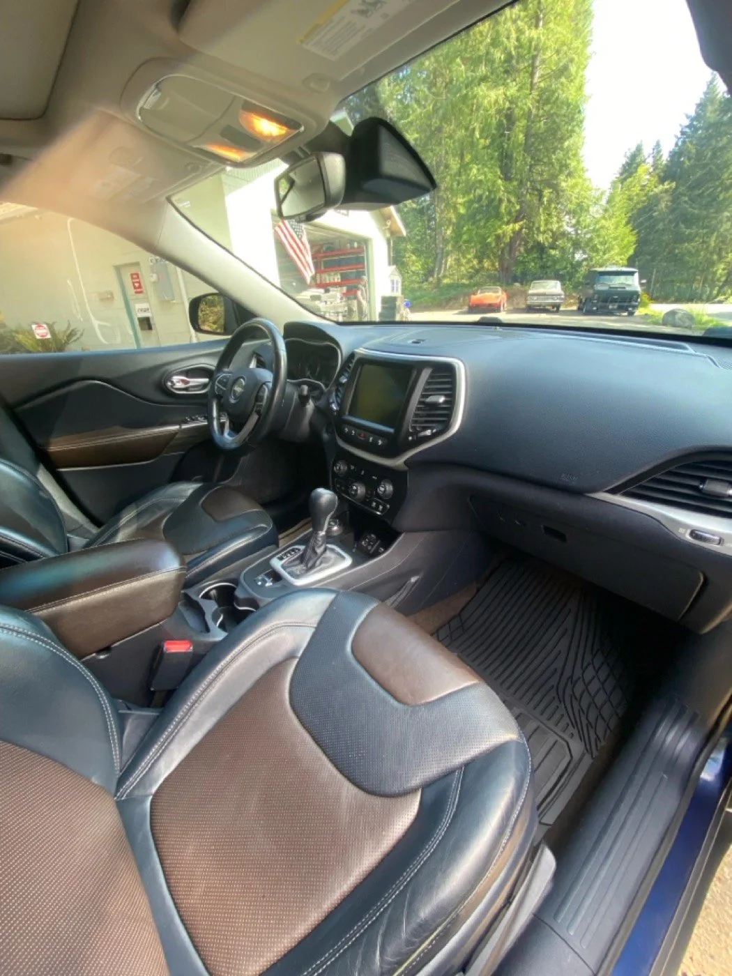 The interior of a vehicle showing the dashboard, steering wheel, gear shift, and front seats with leather upholstery. Outside the vehicle, trees and parked cars are visible.