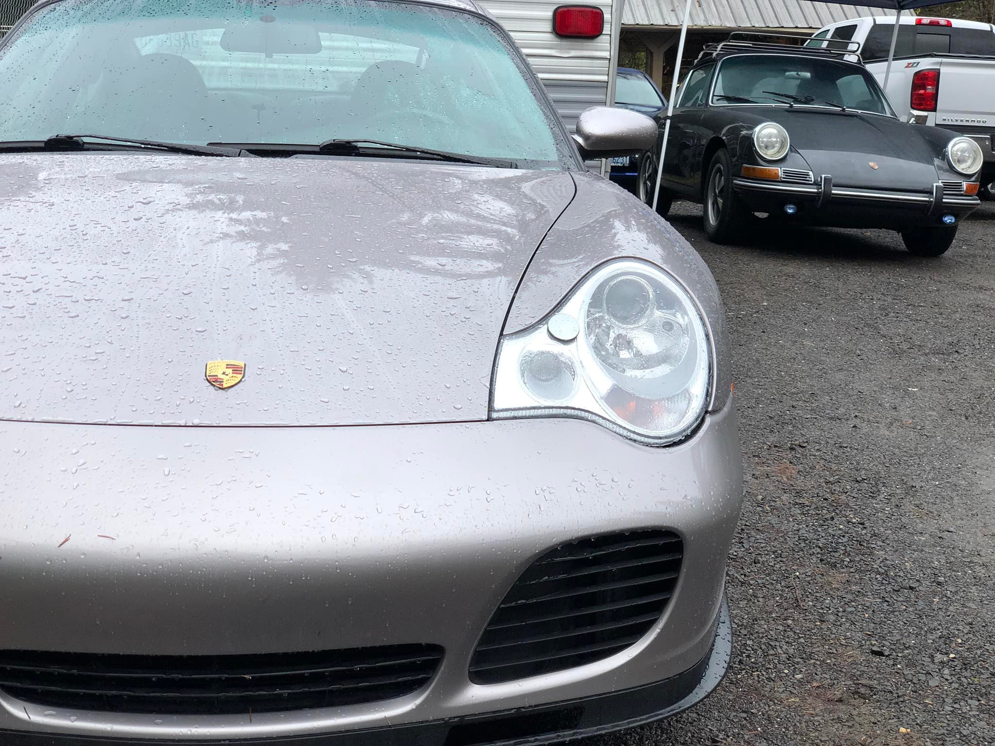 Front of a silver Porsche sports car, with wet raindrops on the hood and windshield, parked in front of other vehicles including a black Porsche and a white truck, in a gravel lot.