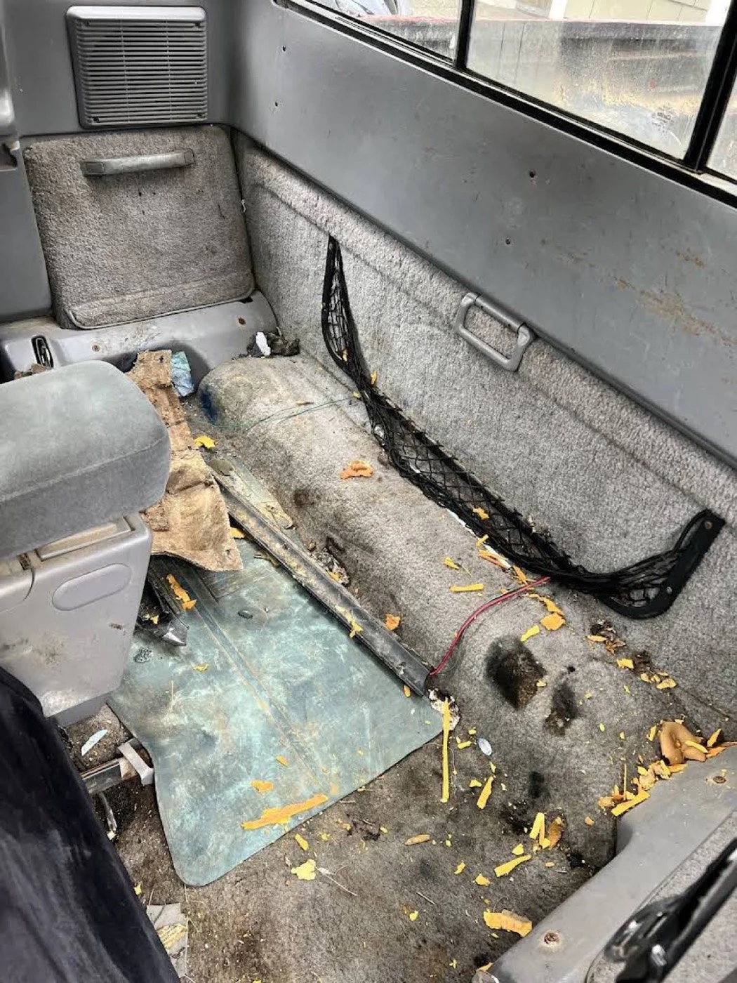 A dirty and damaged interior of a vehicle, with the passenger seat removed, exposing the floor, threshold, and side panels. Debris, yellow leaves, and exposed wires are scattered on the floor. The side panels are worn and torn, and the floor has a pl
