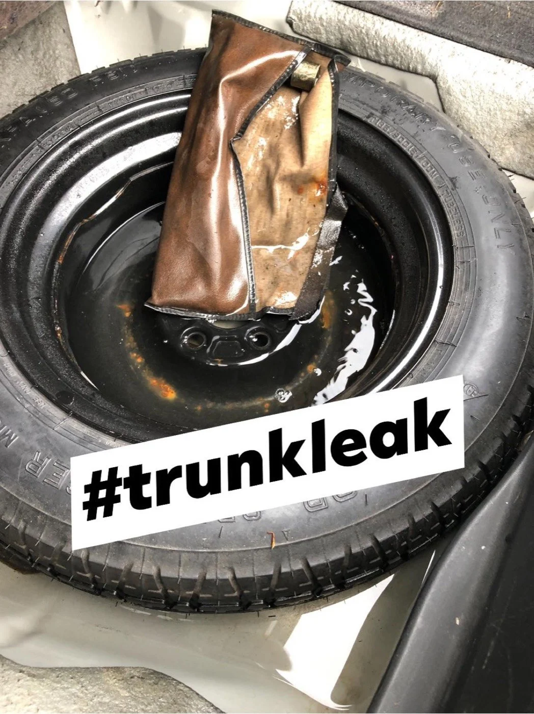 A spare tire with a rusty and damaged trunk liner inside it, with a black liquid at the bottom, and the hashtag '#trunkleak' written across the image.
