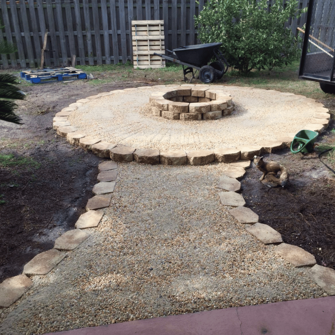 Backyard with a newly built stone fire pit and pathway, surrounded by a wooden fence, with gardening tools and a small dog nearby.