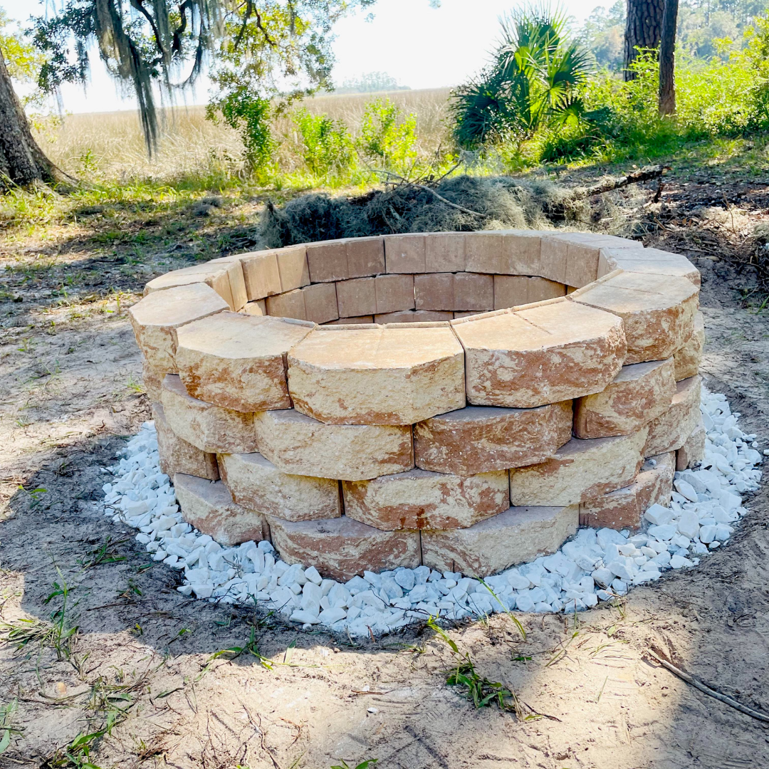 A circular outdoor fire pit made of large beige bricks, surrounded by white gravel, situated on sandy ground in a backyard with trees and greenery in the background.
