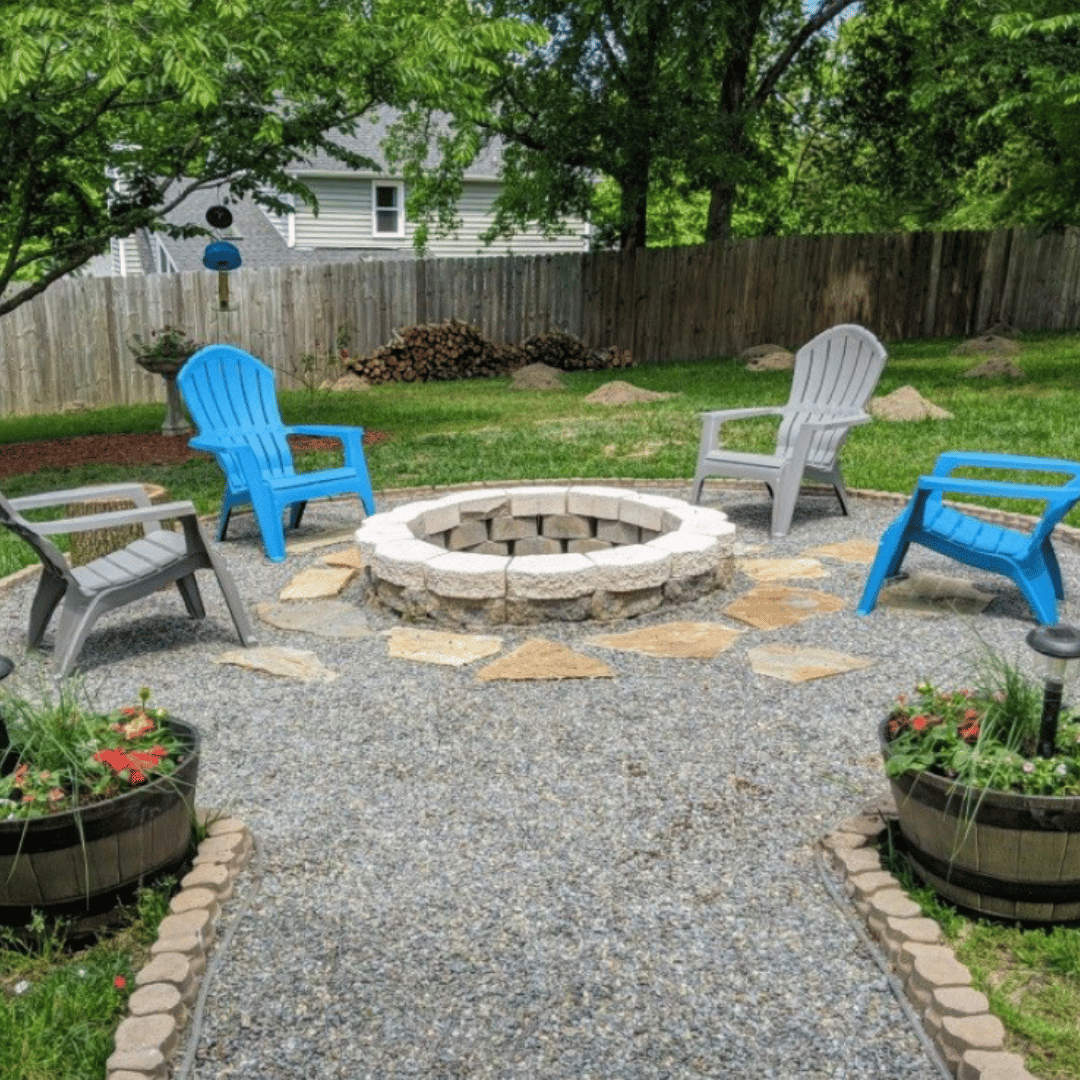 Backyard with a gravel patio, four chairs (two blue, two gray) around a fire pit, potted plants along the pathway, trees, and a wooden fence.