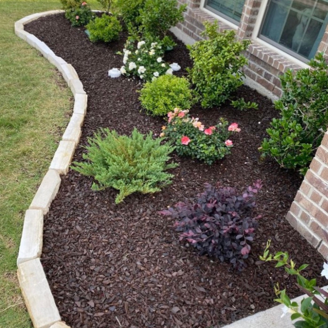 A landscaped garden bed along a brick house, with various green shrubs and flowering plants, bordered by stone edging and filled with dark mulch.