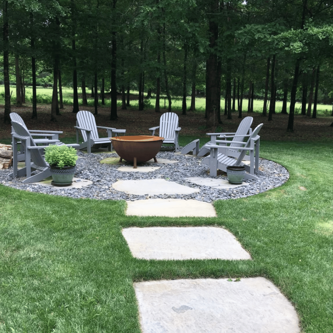 A backyard fire pit area with five gray Adirondack chairs arranged in a circle, two potted plants, and a fire bowl in the center, surrounded by a stone pathway and lawn, with a wooded area in the background.