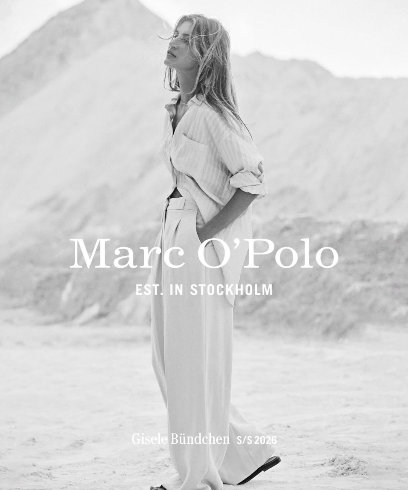 Black and white fashion magazine cover featuring a woman standing in a beach landscape, wearing light-colored wide-leg pants and a loose shirt, with her hands in her pockets and her hair blowing in the wind. Text on the cover reads "Marc O'Polo," "Est. in Stockholm," and "Gisele Bündchen S/S 2026."