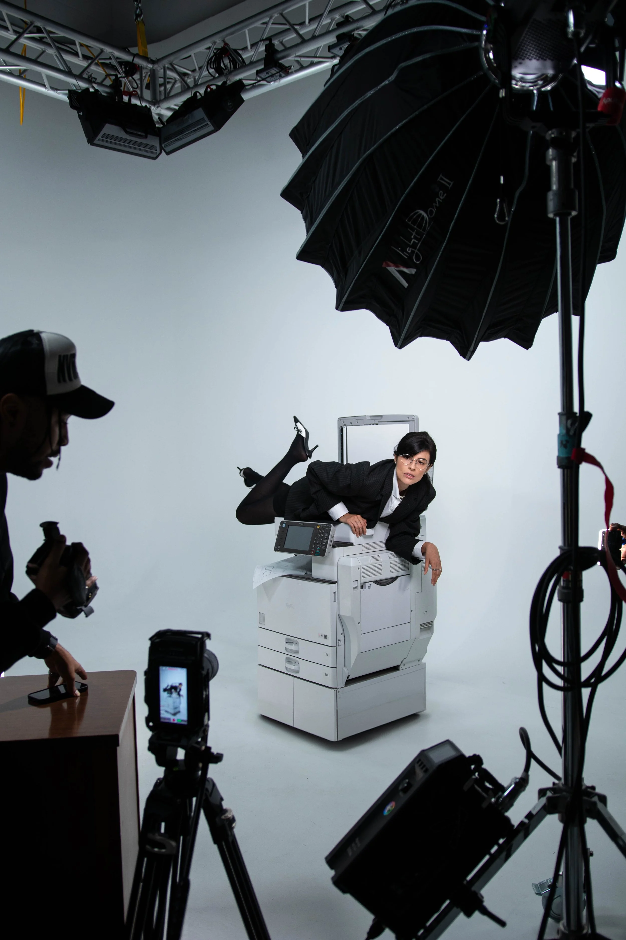 A woman in a suit lying on top of a photocopy machine, with four other people around her taking pictures in a professional photo studio with studio lighting and equipment.