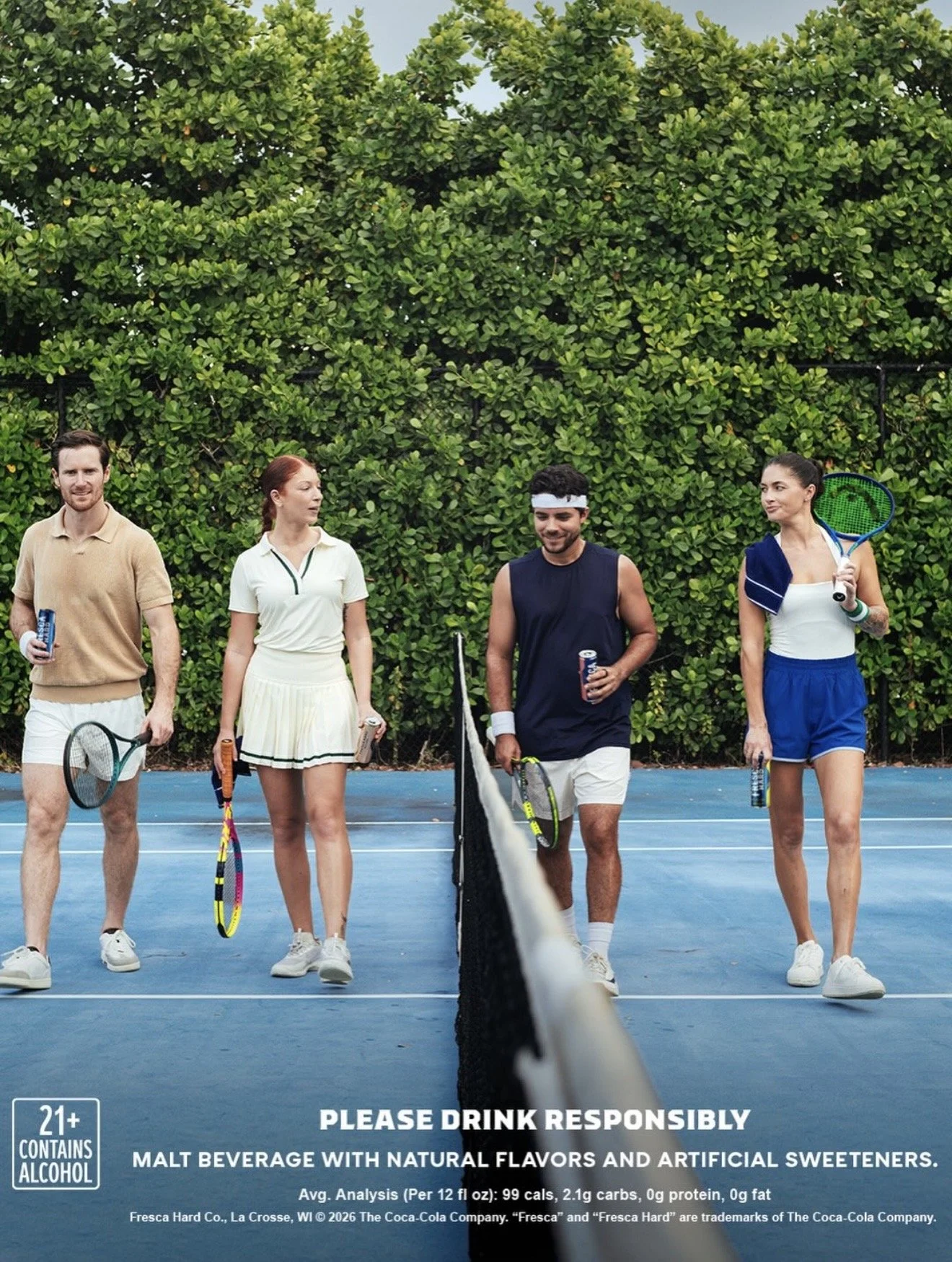 Four people walking on a tennis court holding tennis rackets and drinks after playing tennis, with a green hedge in the background.