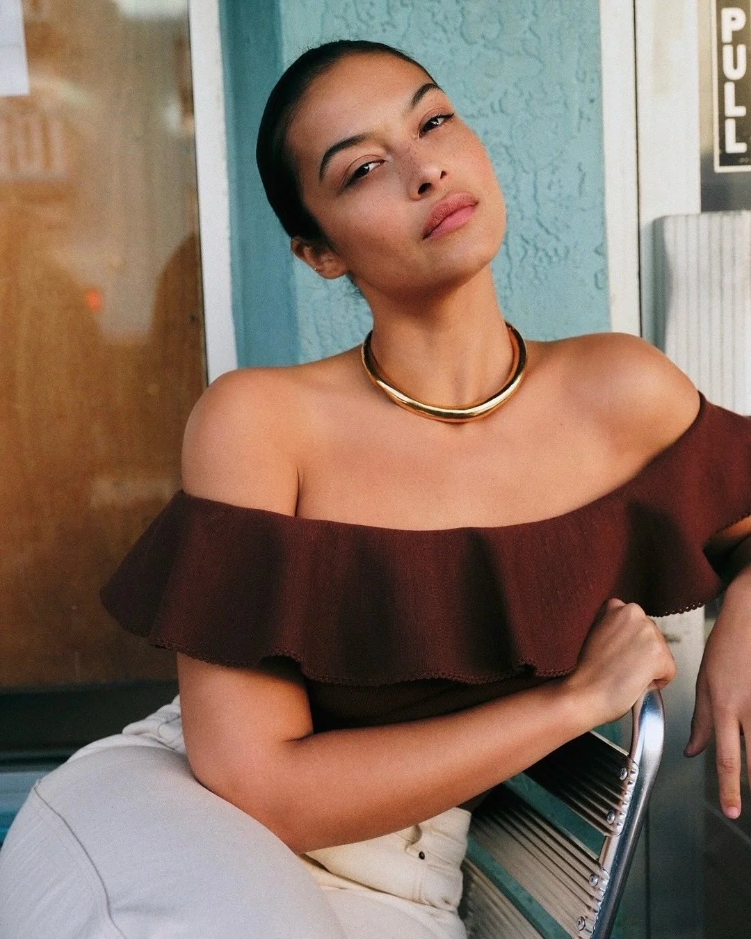 A young woman with dark hair tied back, wearing an off-the-shoulder dark red top and a gold necklace, sitting on a metal bench with a serious expression.