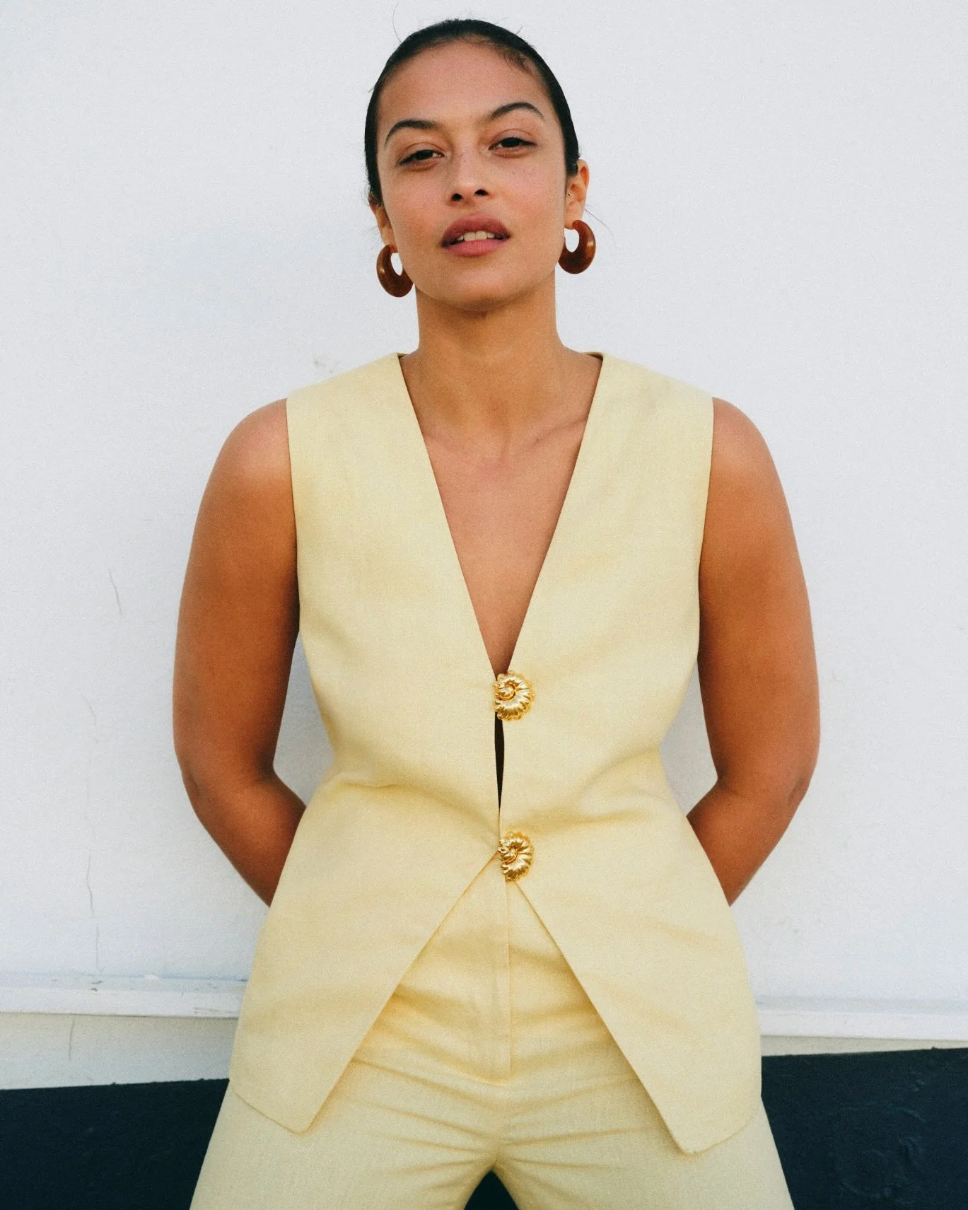 A woman in a sleeveless yellow dress with a V-neckline and gold buttons, standing against a plain white wall. She has dark hair pulled back, wears large hoop earrings, and has a confident expression.