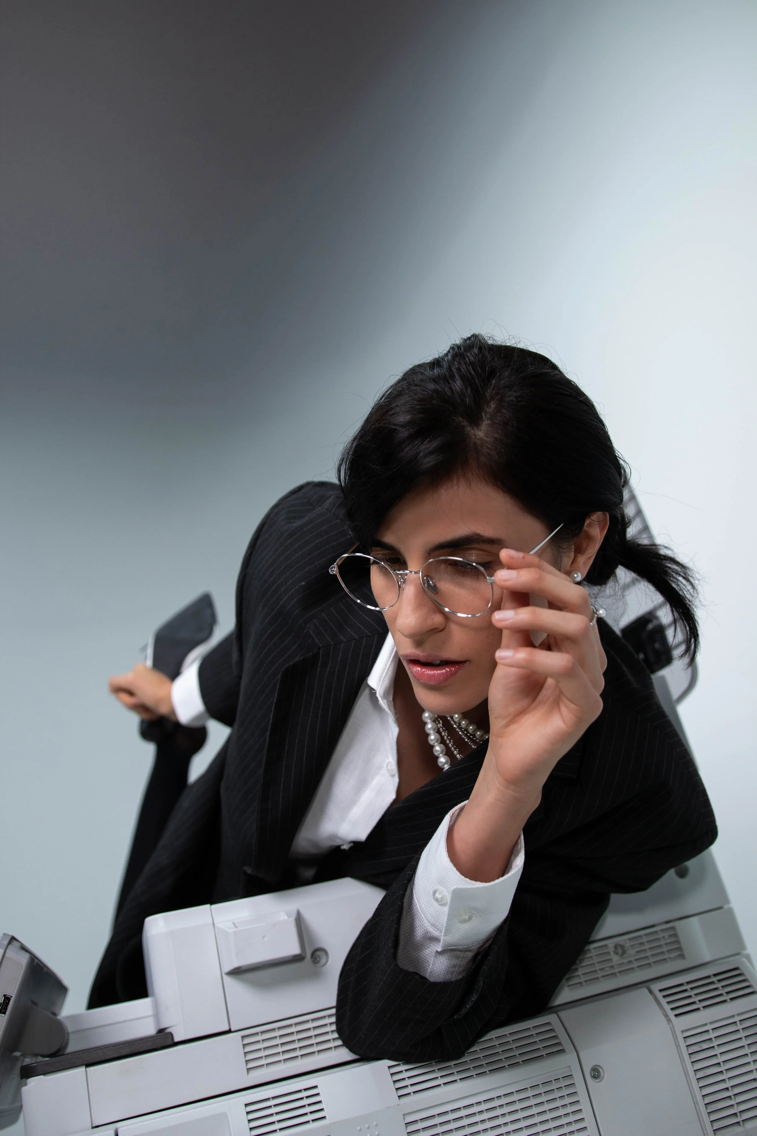 Businesswoman wearing glasses, in a business suit, thinking or concentrating, with printers or copiers stacked in front of her.