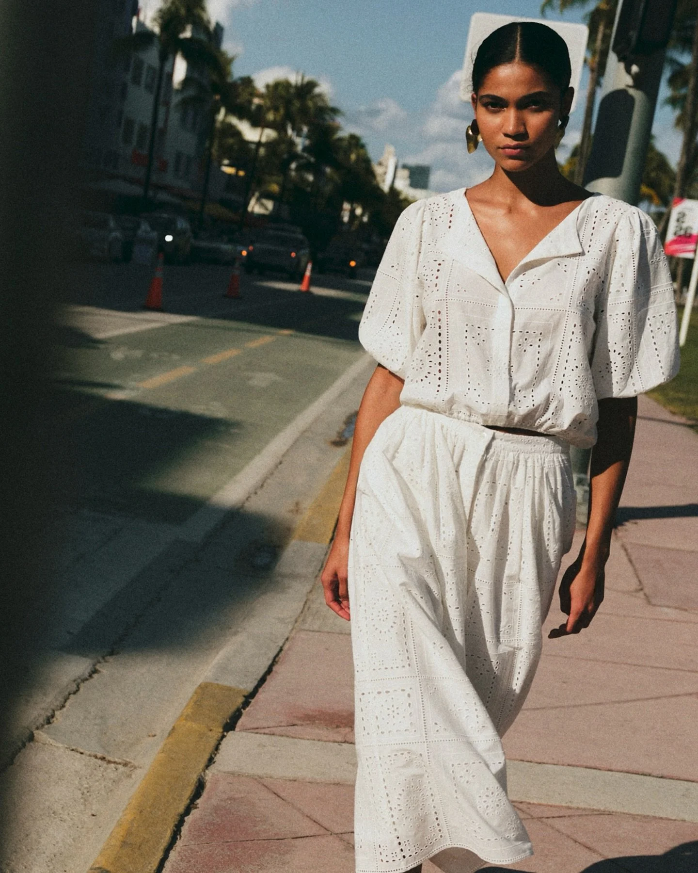 A woman walking on the sidewalk wearing a white eyelet dress with puffed sleeves, black hair pulled back, gold earrings, holding a smartphone, with cars parked along the street and palm trees in the background.