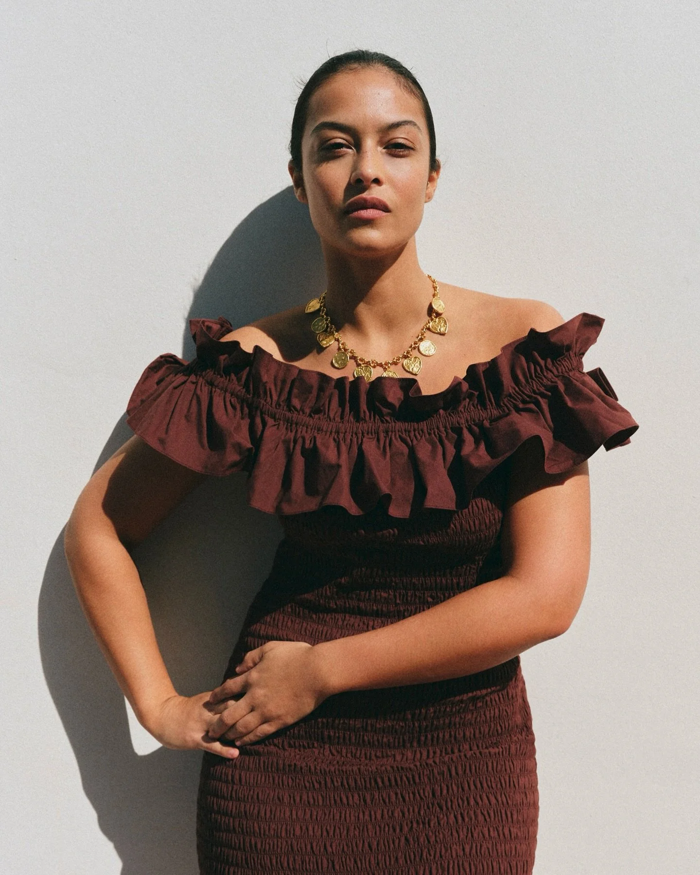 A woman with dark hair pulled back, wearing a ruffled off-shoulder burgundy dress and a gold coin necklace, stands against a plain white wall with her arm on her hip, casting a shadow.