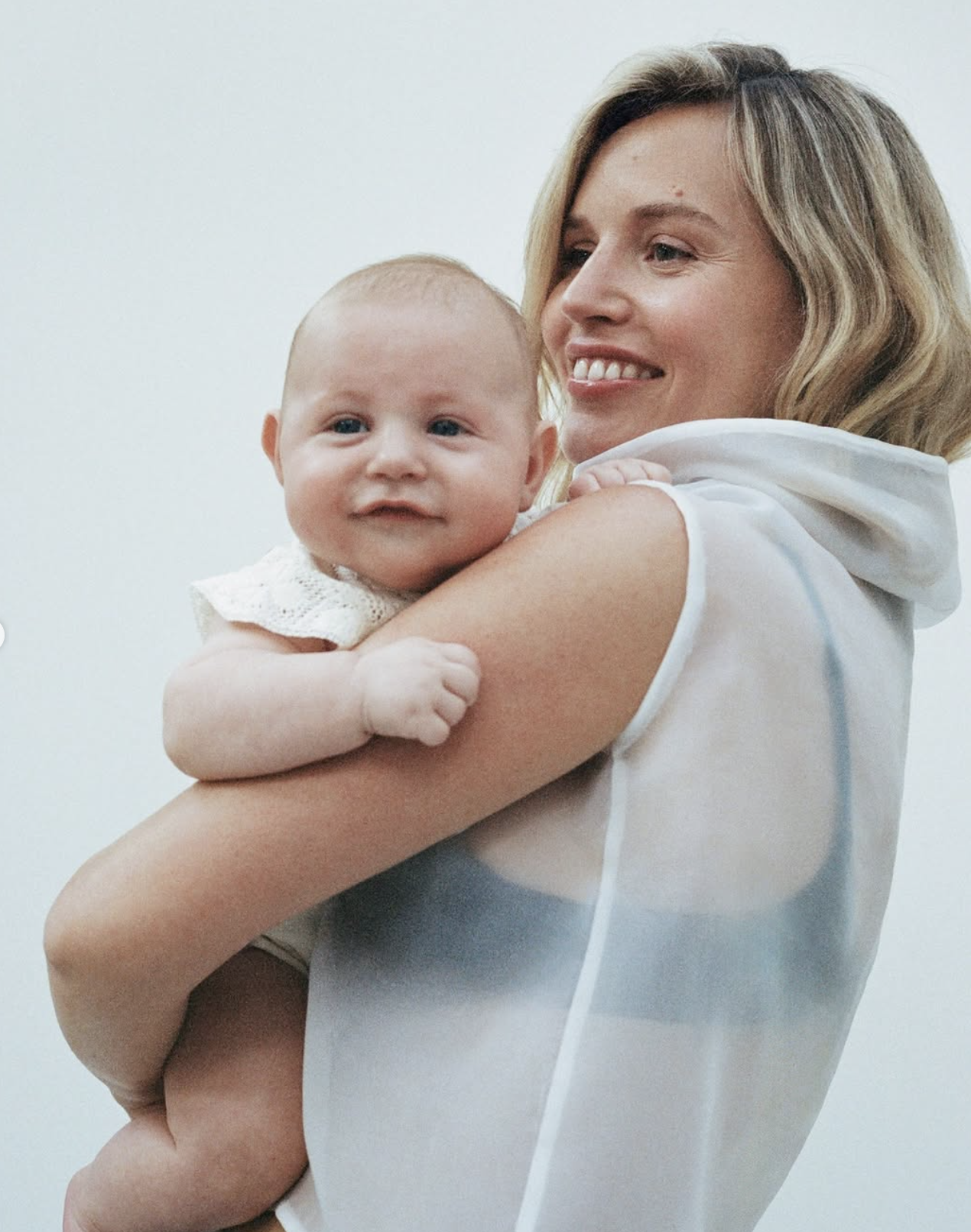 A woman with blonde hair holding a smiling baby with light skin and blue eyes, both dressed in white, against a plain light background.