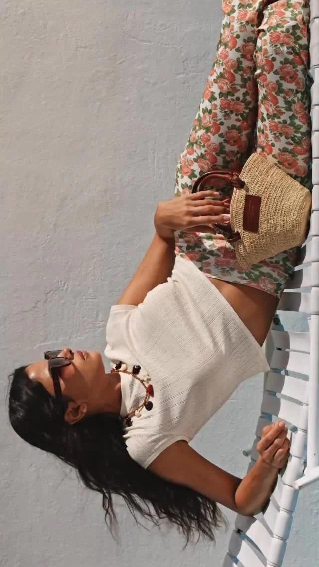 Woman lying on a lounge chair, wearing floral pants, a white top, sunglasses, and a necklace, holding a woven handbag.