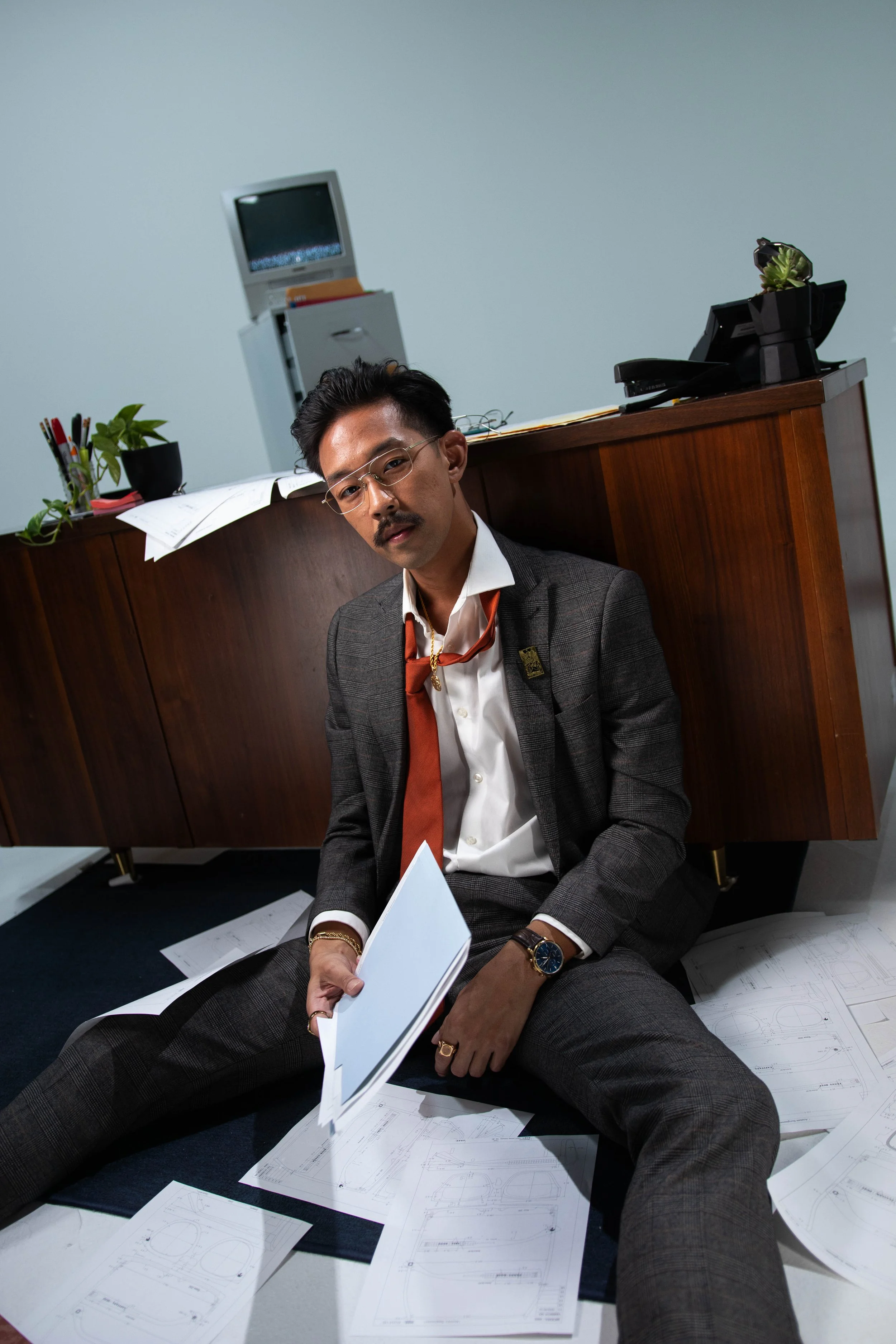 A man with glasses and a mustache sitting on the floor in an office, surrounded by scattered blueprints, holding a folder, wearing a suit and tie, with a disorganized desk and papers behind him.