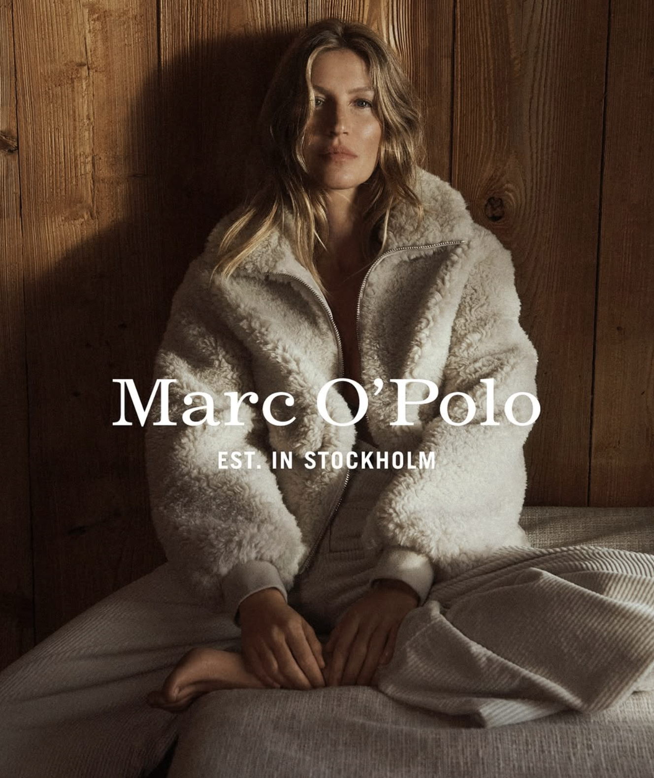 A woman sitting on a bed against a wooden wall, wearing a fluffy zippered jacket. The text overlay reads "Marc O'Polo EST. IN STOCKHOLM."