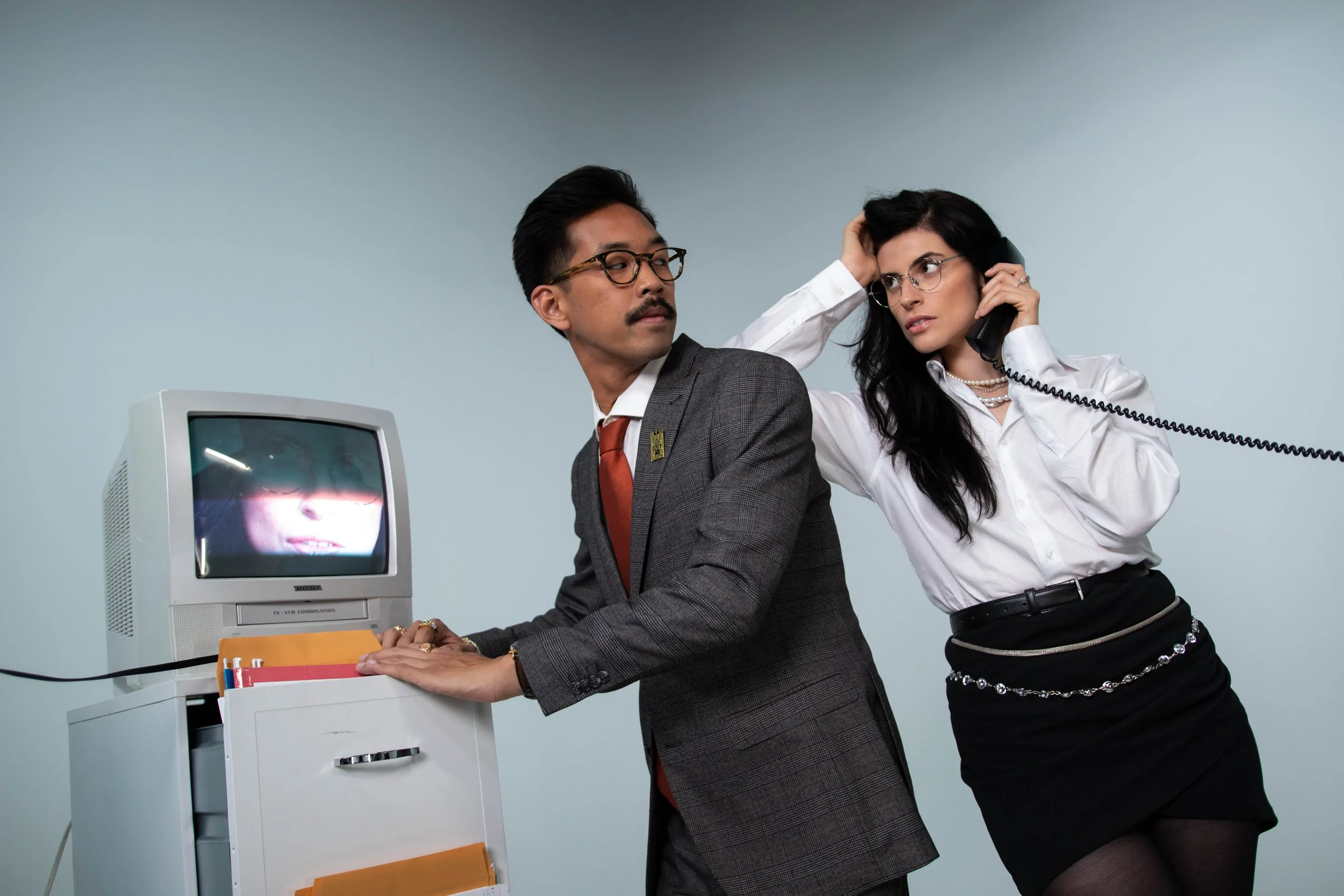 A man with glasses and a mustache wearing a suit sits at a desk, using a vintage computer. A woman with glasses, dressed in a white shirt and black skirt, is holding a phone to her ear and instinctively touches her head.