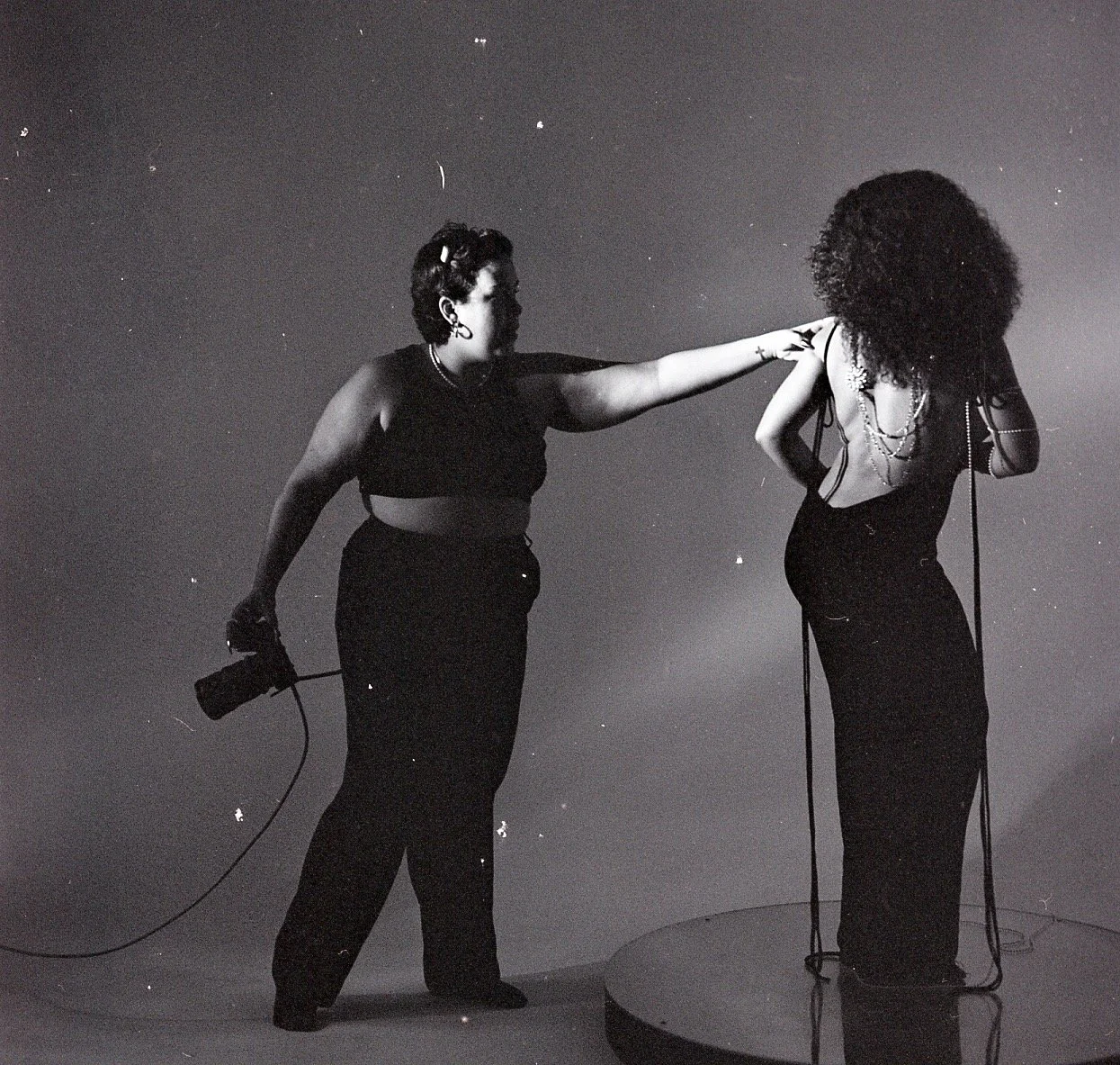 Two women are in a photo, one holding a camera, with her arm extended towards the other woman, who has her head down and her face obscured by curly hair. The scene appears to be in a studio setting with a plain background.