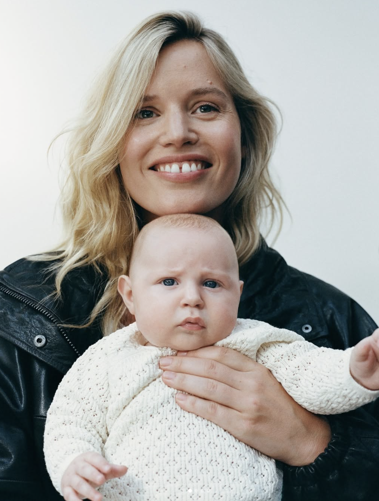 A smiling woman with blonde hair holding a serious-looking baby with light hair, wearing a cream-colored sweater.