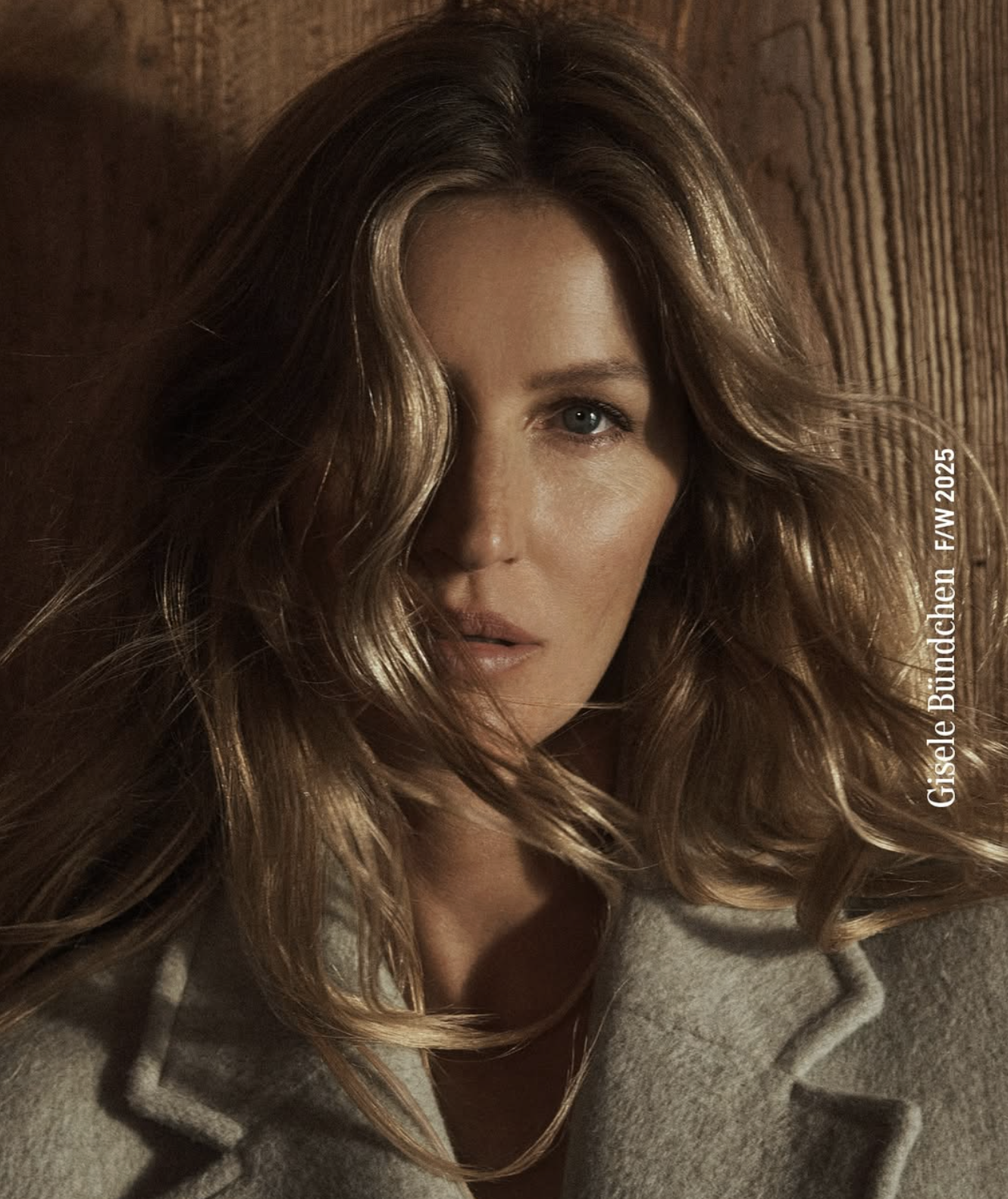Close-up of a woman with wavy, light brown hair, blue eyes, and natural makeup, wearing a gray hoodie, leaning against a wooden wall. Text overlay reads 'Giselle Bundchen F/W 2025'.