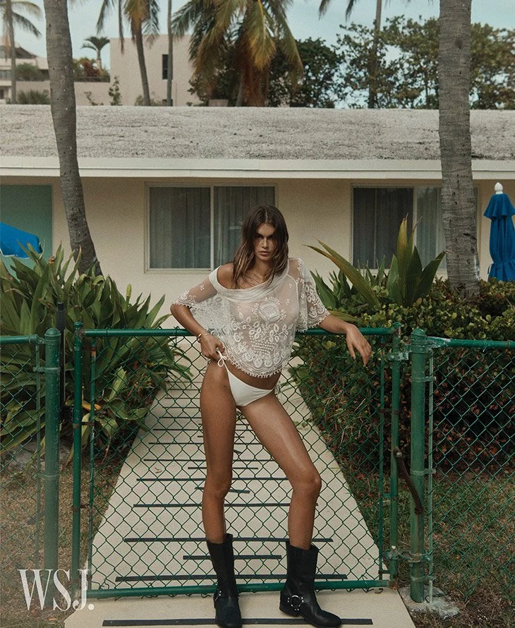 A woman with wavy brown hair standing on a small bridge or walkway with a chain-link fence, in front of a beige building with large windows, palm trees, and tropical plants. She is wearing a sheer lace top, white underwear, and black boots, posing confidently.